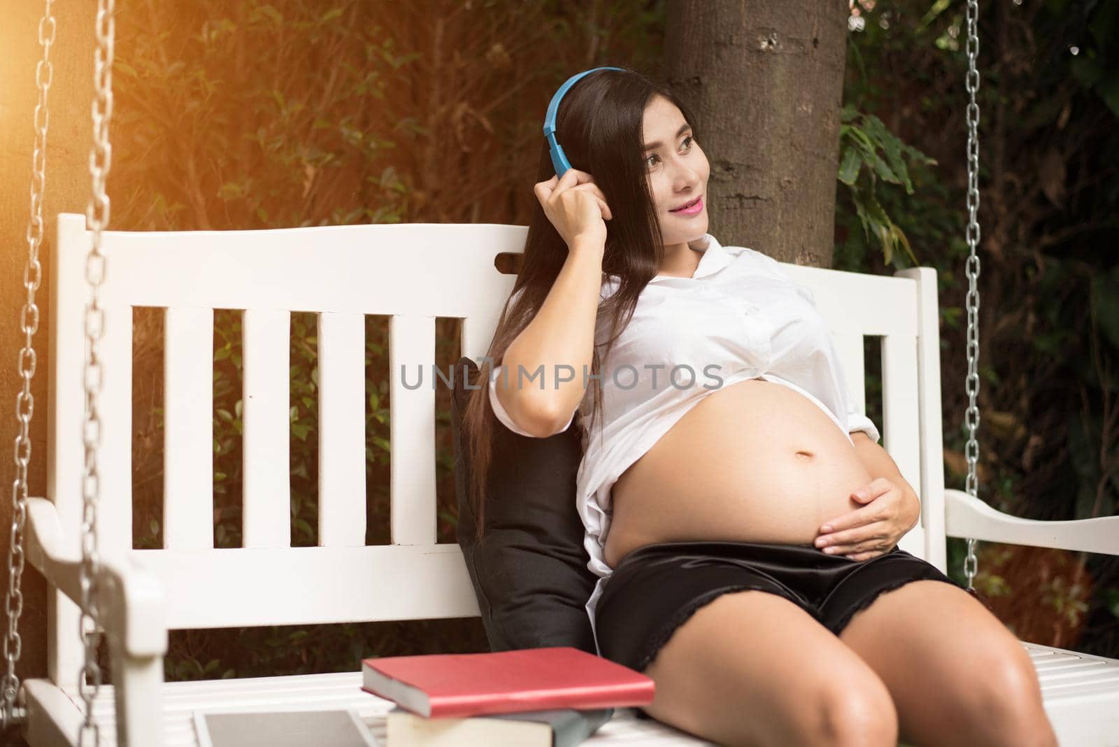 Asian young pregnant woman relaxing and listening music on swing in park garden with books. People and Entertainment concept. Happiness and pleasure concept. Single mom and Widow theme. 