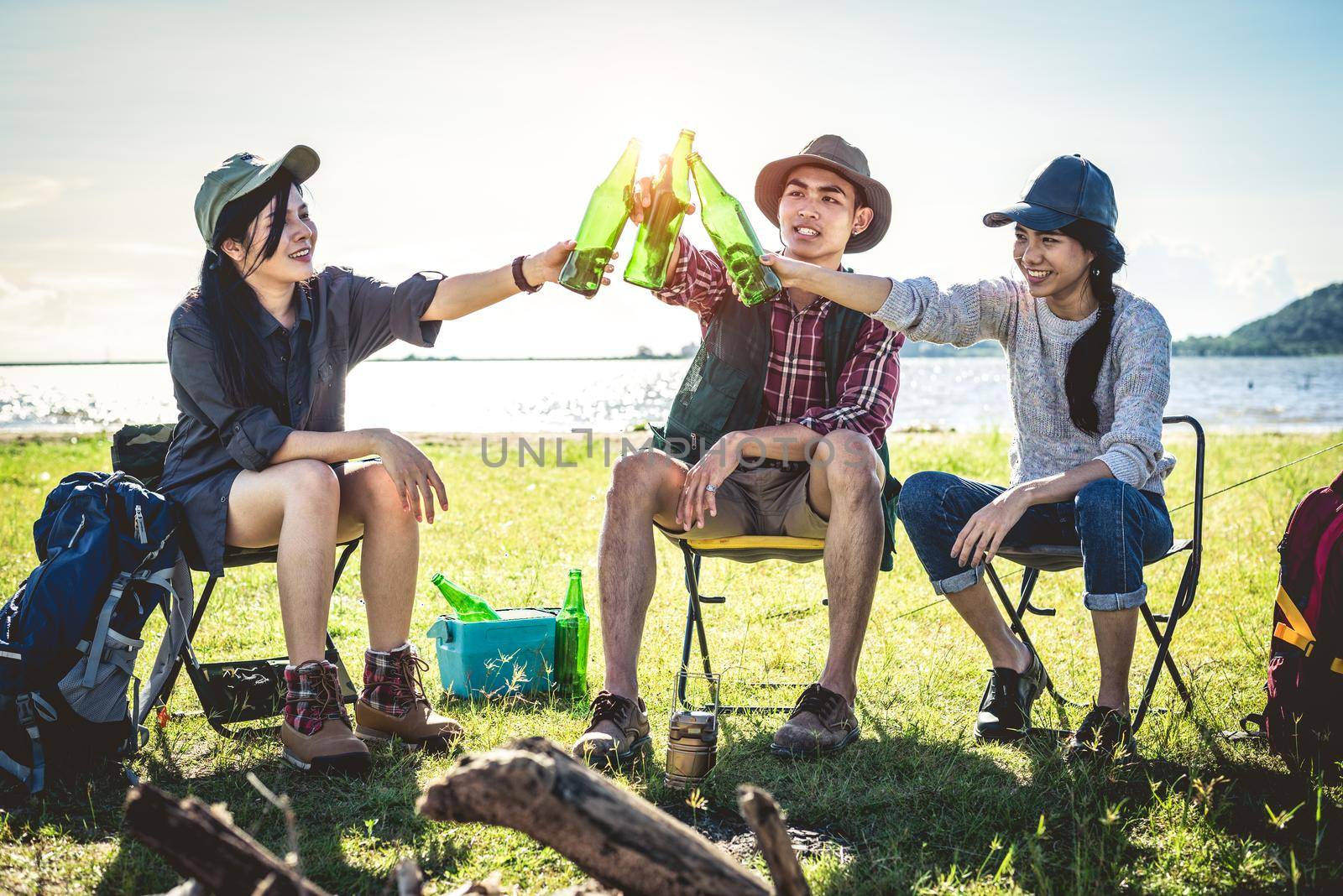 Group of young Asian friends enjoy picnic and party at lake with camping backpack and chair. Young people toasting and cheering bottles of beer. People and lifestyles concept. Outdoor background theme by MiniStocker