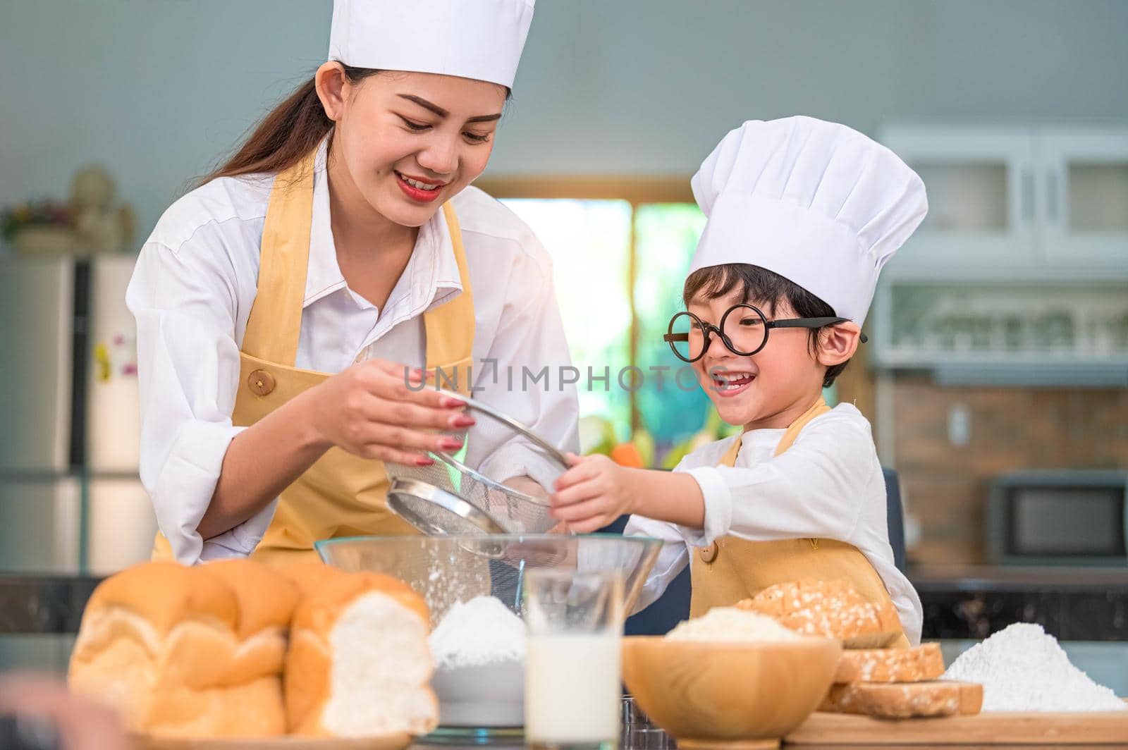 Cute little Asian boy and beautiful mother sifting dough flour with sifter sieve colander in home kitchen on table for prepare to baking bakery and cake. Thai kids playing with flour as chef funny