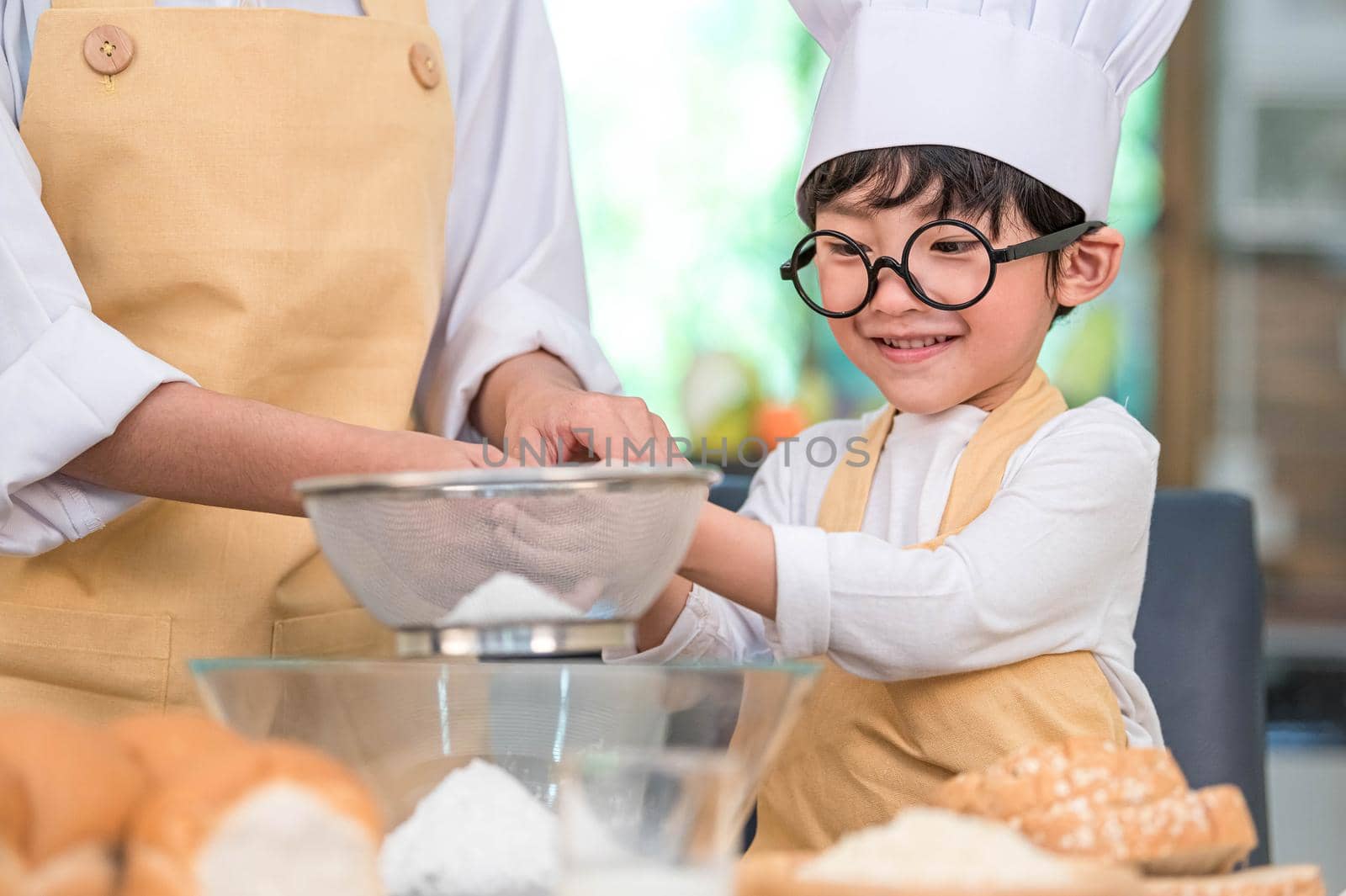 Cute little Asian boy and beautiful mother sifting dough flour with sifter sieve colander in home kitchen on table for prepare to baking bakery and cake. Thai kids playing with flour as chef funny