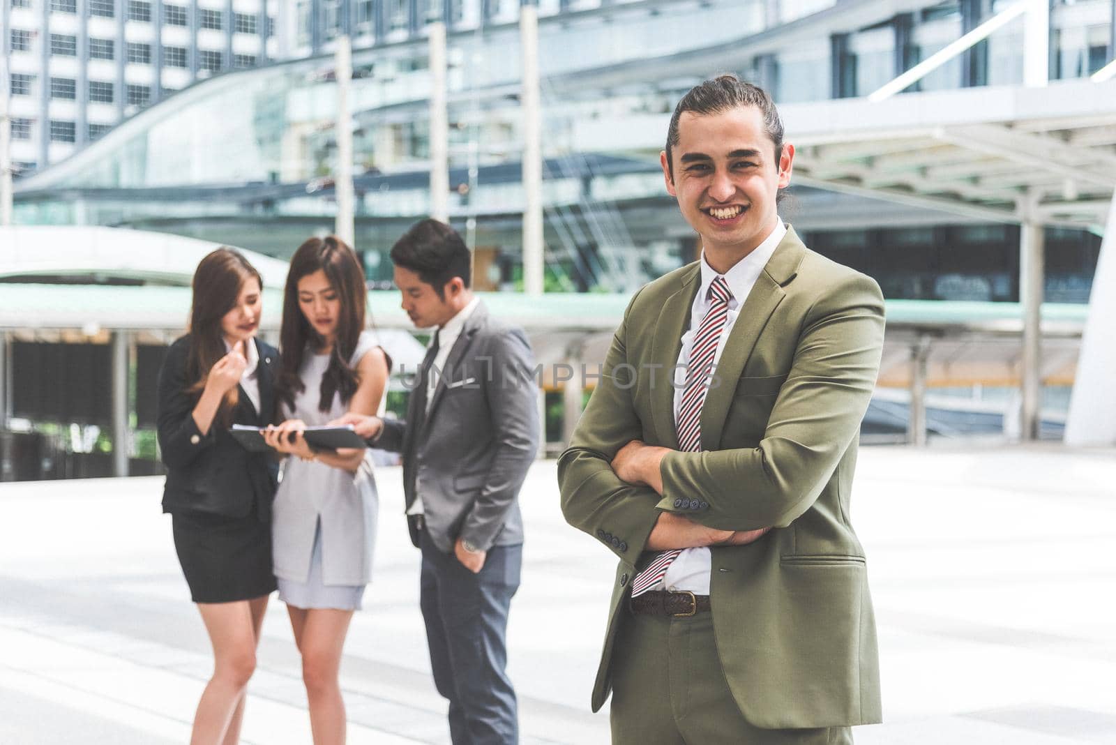 Smiling young businessman looking into camera with his arms crossed and teammates background in urban. Modern business office team at outdoors in city. Business people and Leadership lifestyle concept by MiniStocker