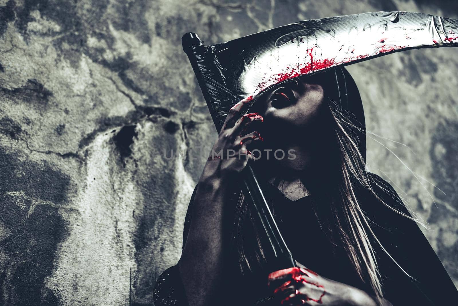 Witch licking blood on reaper. Female demon angel in black clothes and hood on grunge wall background. Halloween day and Mystery concept. Fantasy of magic theme. Afterlife and Death concept.