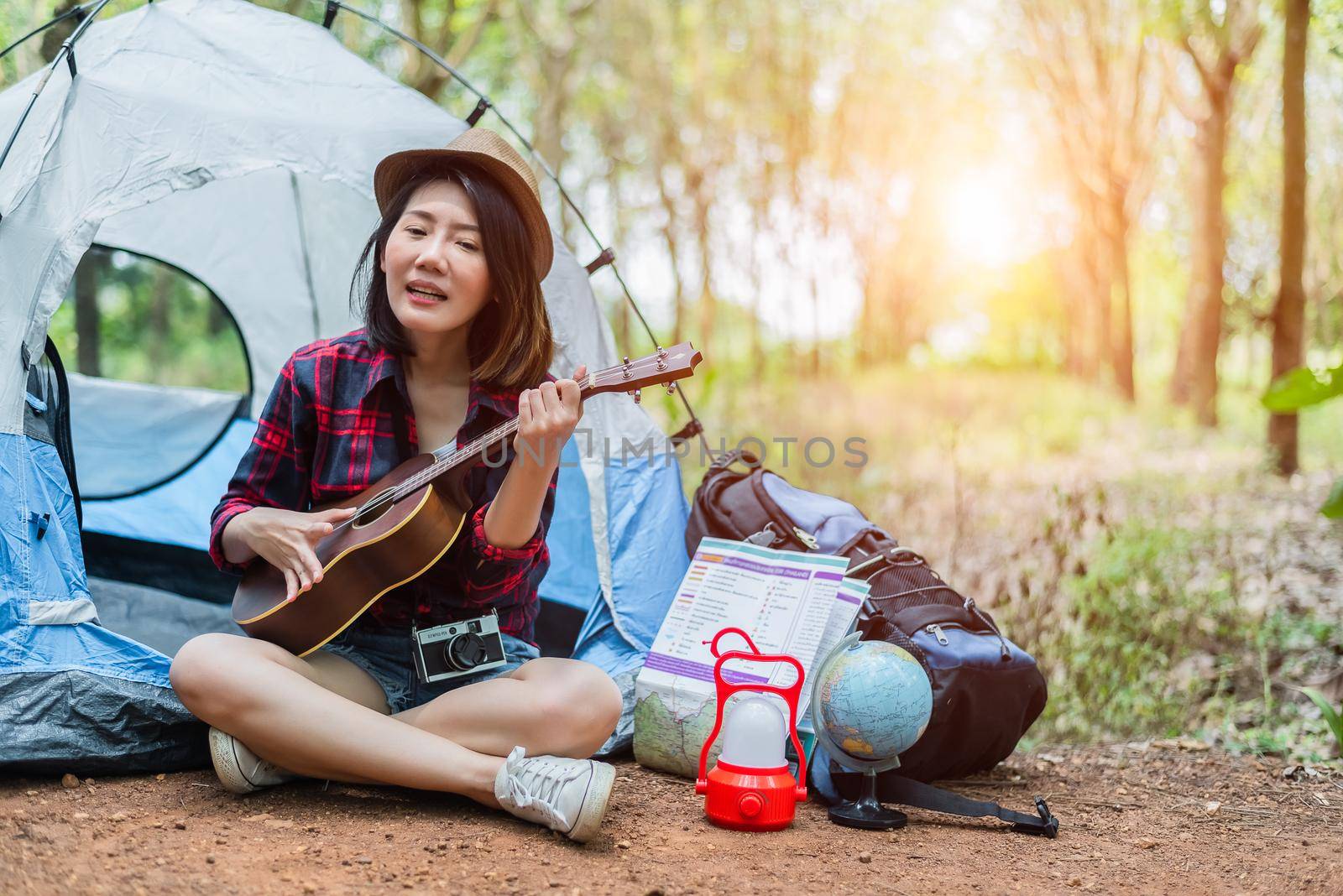 Beautiful Asian woman playing Ukulele in front of camping tent in pine woods. People and Lifestyles concept. Adventure and Travel theme.