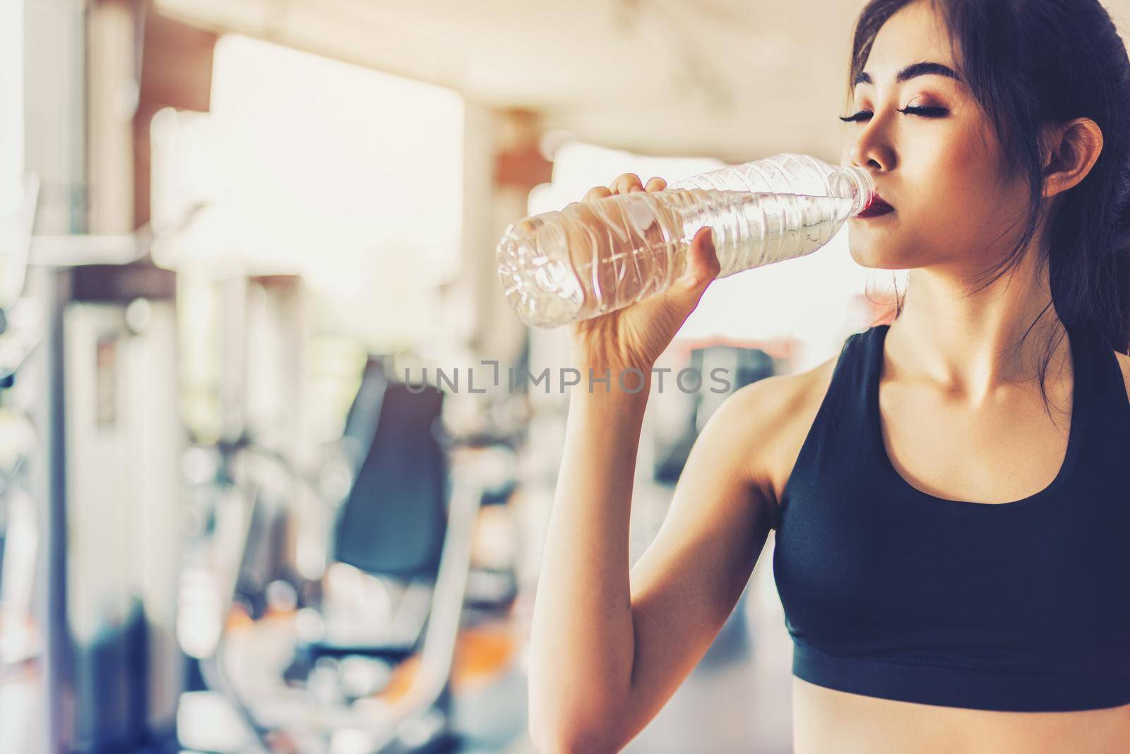 Asian woman drinking pure drinking water for freshness after workout or exercise training in fitness gym with fitness equipment background. Relax and Rest concept. Strength and Fitness training theme.