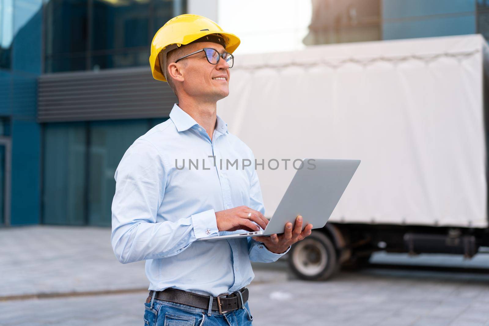 Business. Engineer Worker Protective Helmet Use Laptop Controls Working Process Inspector Supervisor Yellow Hard Hat Glasses Transportation Company Office Building And Truck Background