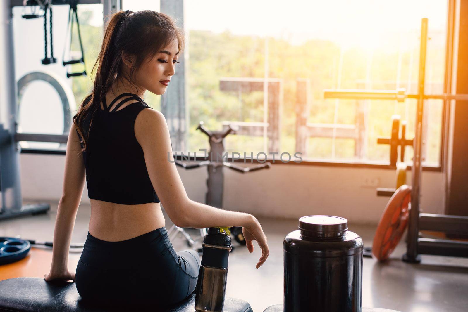 Sport woman sitting and resting after workout or exercise in fitness gym with protein shake or drinking water on floor. Relax concept. Strength training and Body build up theme. Warm and cool tone