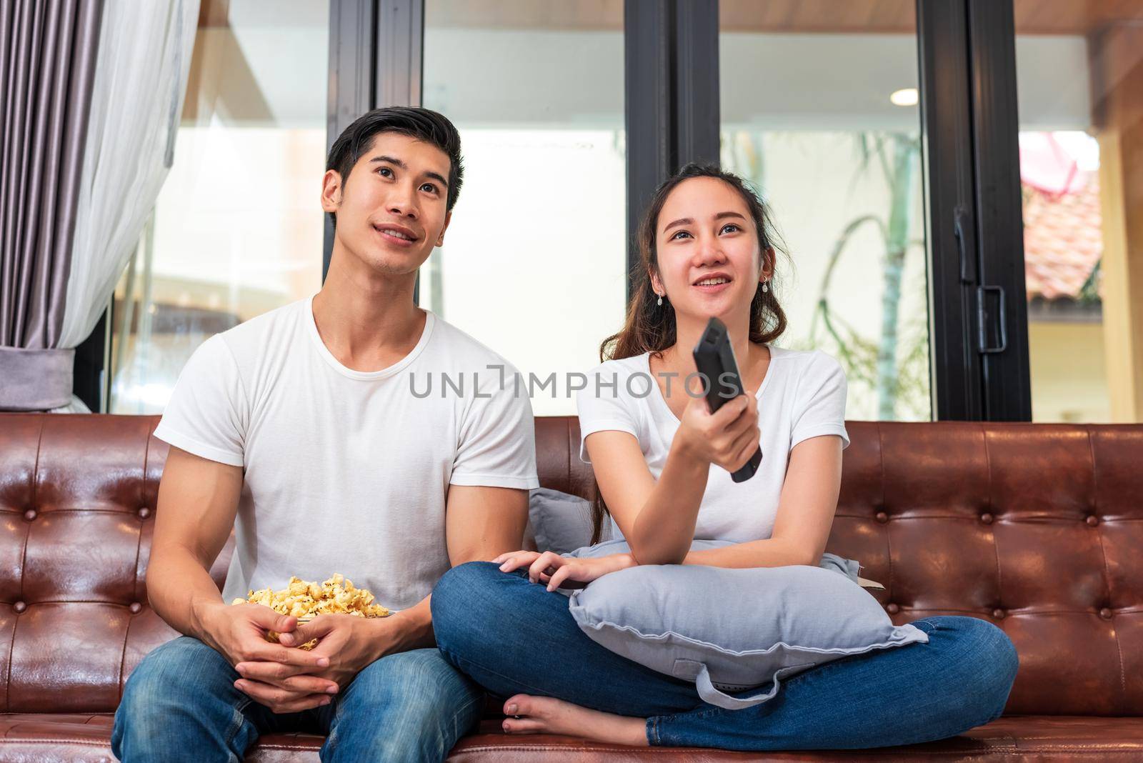 Asian couples watching television together on sofa in their home. People and lifestyles concept. Vacation and holiday concept. Honeymoon and pre wedding theme. Happy family activity in valentines day by MiniStocker