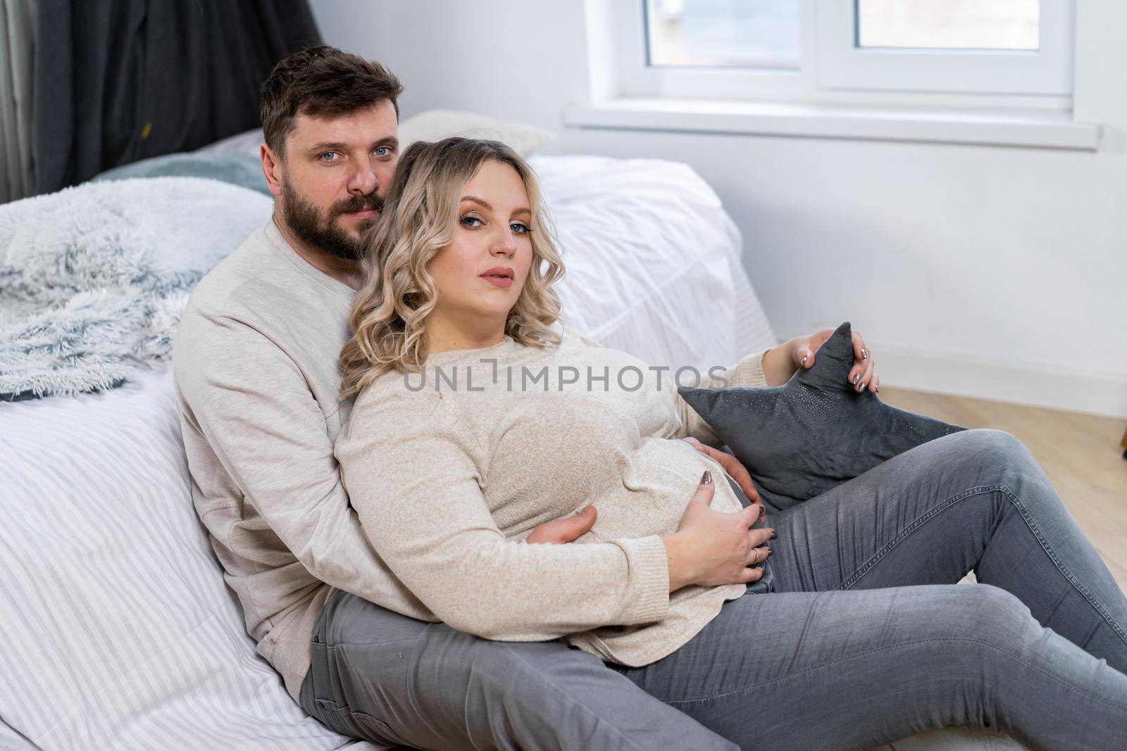 Happy family concept. Husband hug belly pregnant wife sitting floor indoor living room near sofa by andreonegin