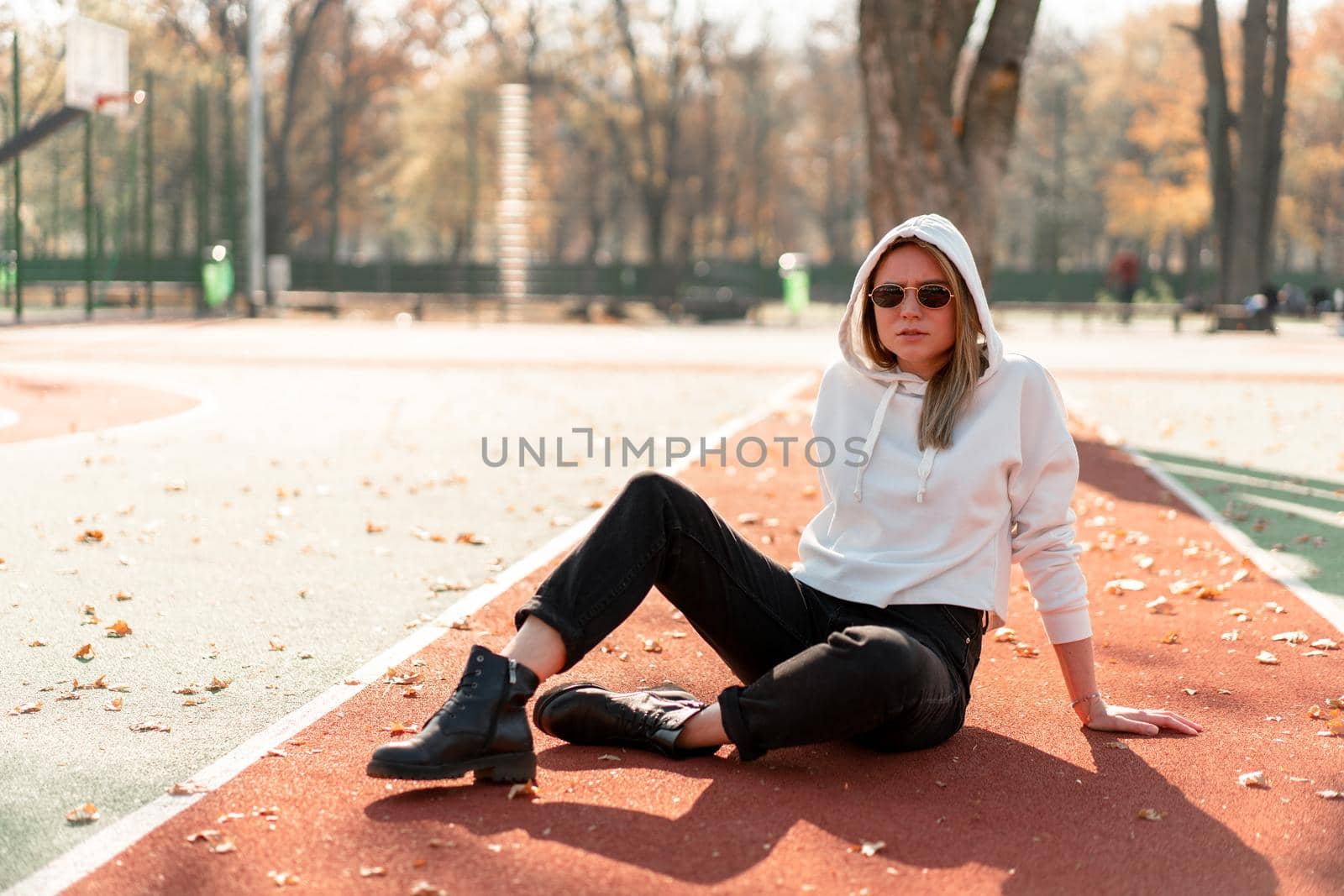 Outdoor portrait of young beautiful woman with long in sunglasses and a white hooded sweater sitting on the sportsground track. youth culture summer pastime