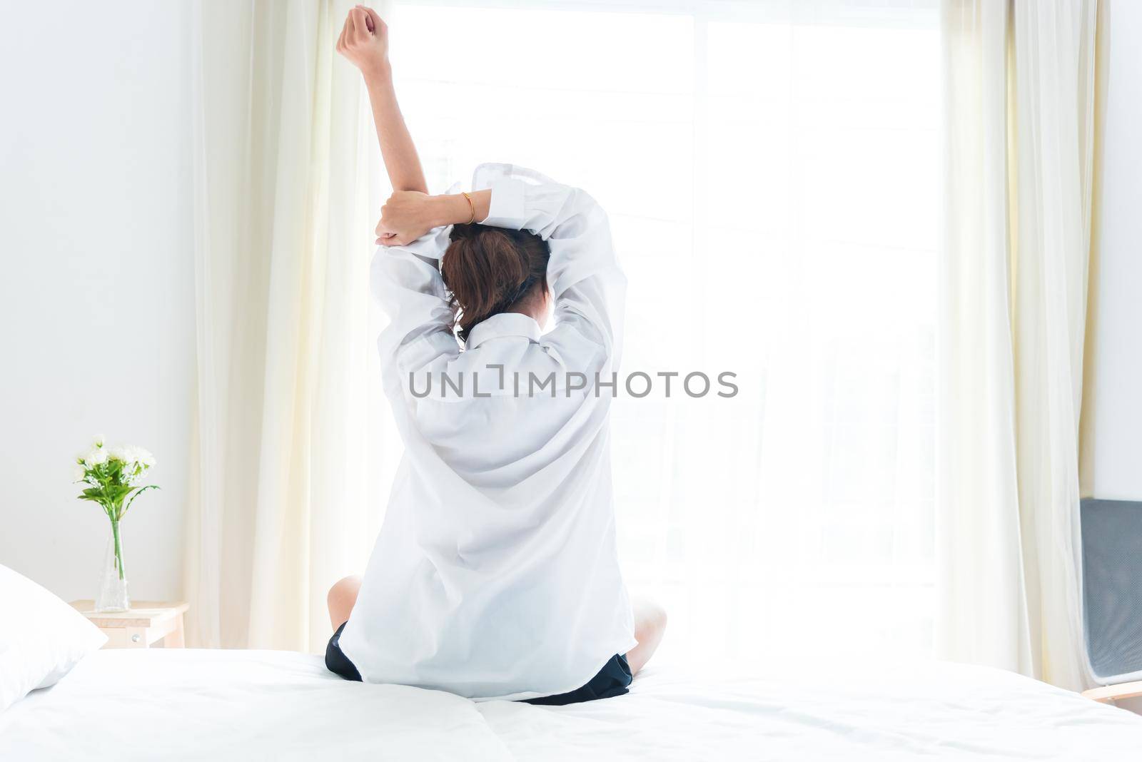 Back view of woman stretching in morning after waking up on bed near window. Holiday and Relax concept. Lazy day and Working day concept. Office woman and worker in daily life theme