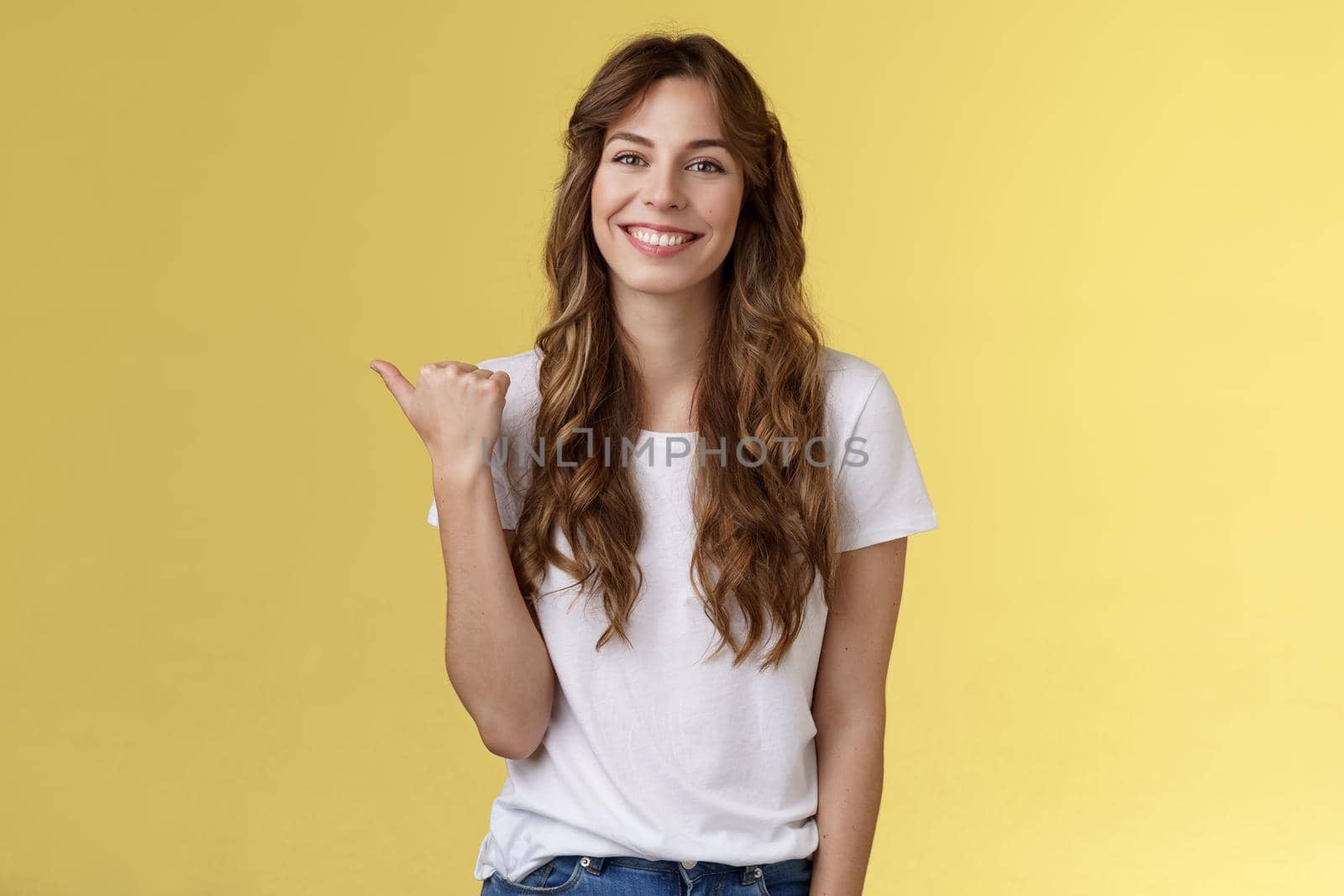 Visit see yourself. Cheerful charismatic good-looking outgoing girl long curly haircut showing place do good hairstyle smiling happily delighted pointing thumb left introduce promo yellow background by Benzoix