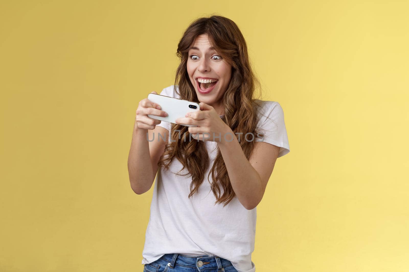 Happy upbeat playful enthusiastic curly-haired girl tempting playing stunning awesome smartphone game hold mobile phone horizontal way smiling broadly stare camera focused winning beat score by Benzoix