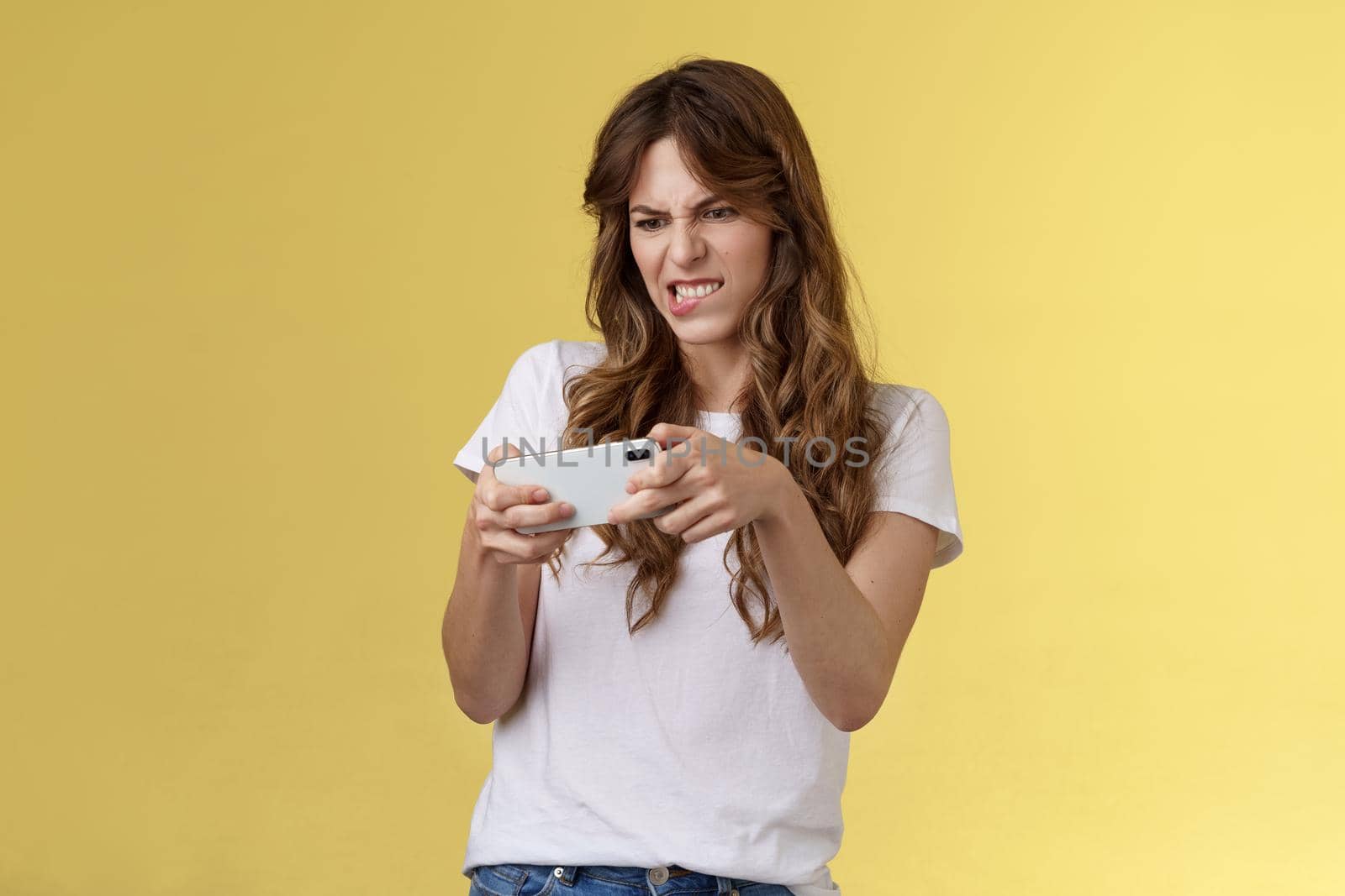 Excited focused playful enthusiastic geeky attractive girl trying beat score hold smartphone horizontal grimacing intense look screen playing awesome arcade game yellow background.