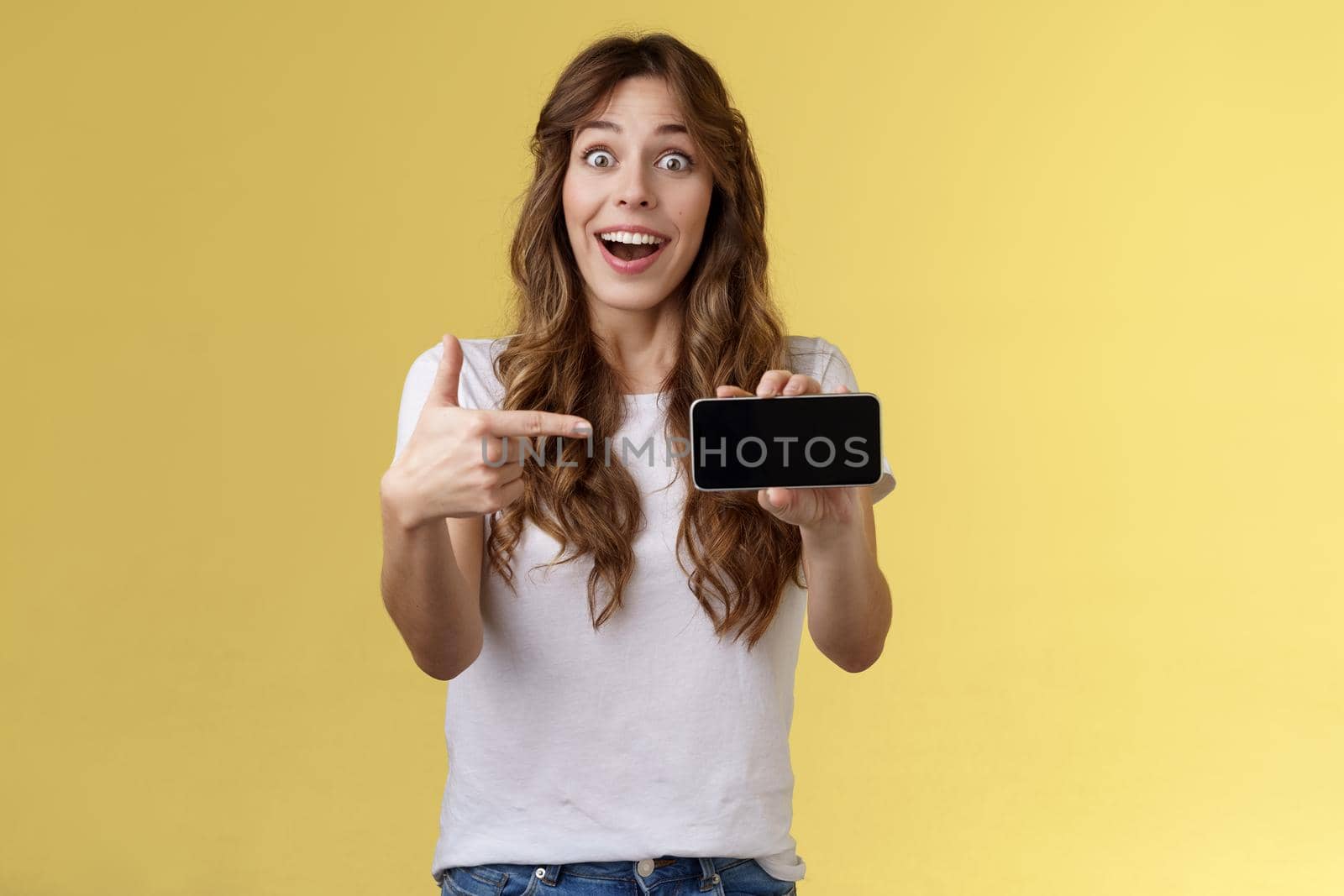 Impressed upbeat happy lucky girl curly long hairstyle open mouth admiration joy like awesome new app show smartphone screen horizontal phone display stand yellow background amazed by Benzoix