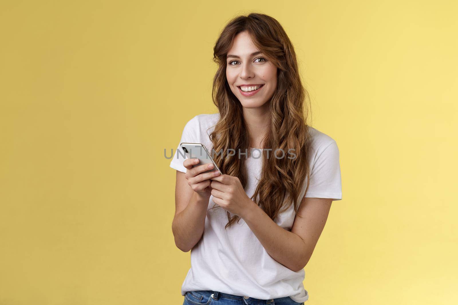 Enthusiastic good-looking urban girl wear white t-shirt standing casual smiling delighted camera texting hold smartphone scroll social media feed stand yellow background browsing network. Lifestyle.