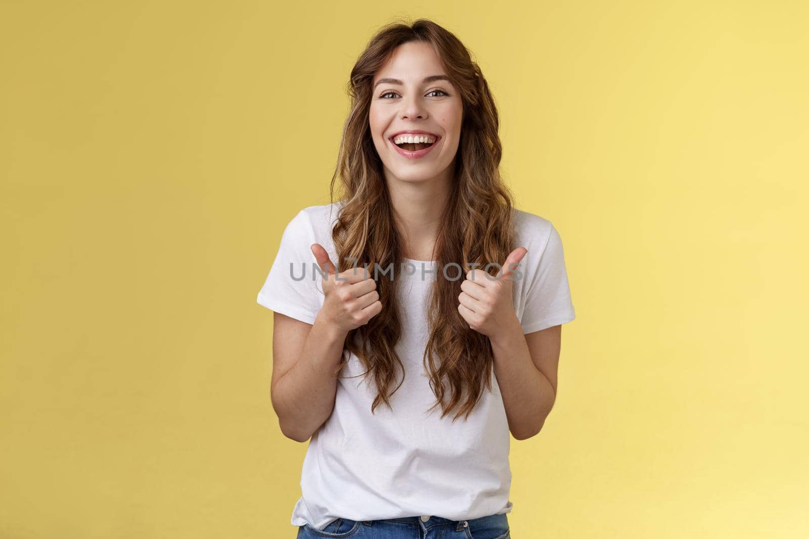 Pleased outgoing cheerful good-looking caucasian girl curly long haircut white t-shirt show thumbs up smiling lively pleased like awesome performance approve good choice yellow background. Lifestyle.