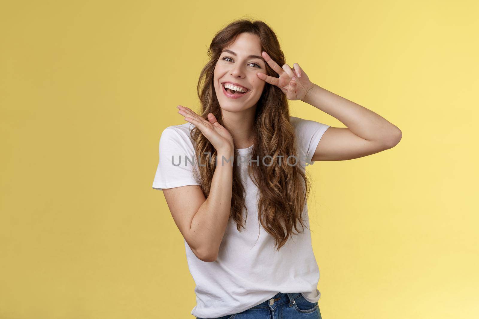 Lively enthusiastic attractive carefree woman enjoy relaxed weekends summer holiday have fun smiling broadly tilt head show peace victory sign joyfully introduce herself posing yellow background. Lifestyle.