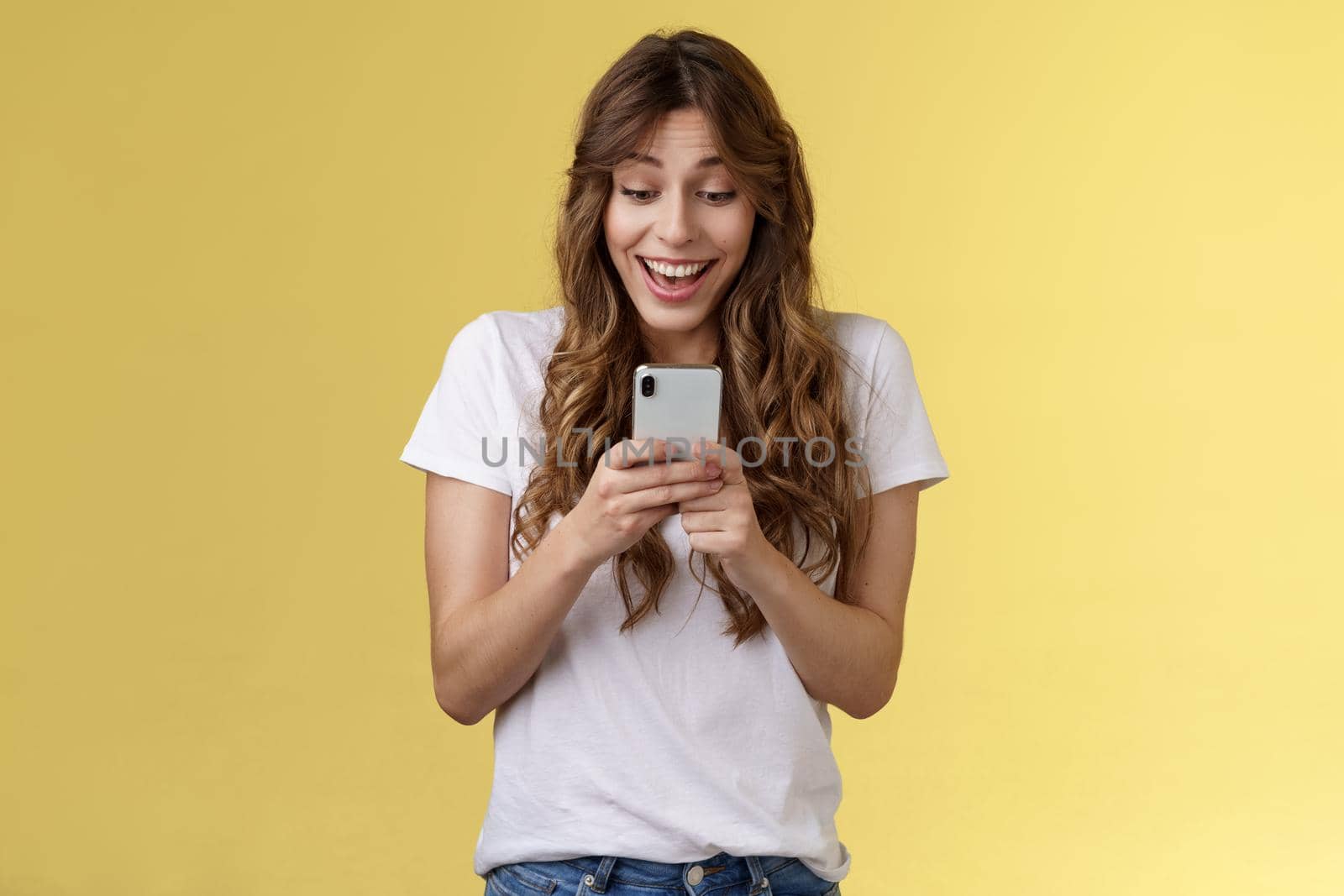 Excited cheerful happy curly-haired silly girl receive surprising good news hold smartphone smiling delighted look mobile phone screen answering b-day invitation stand yellow background upbeat by Benzoix