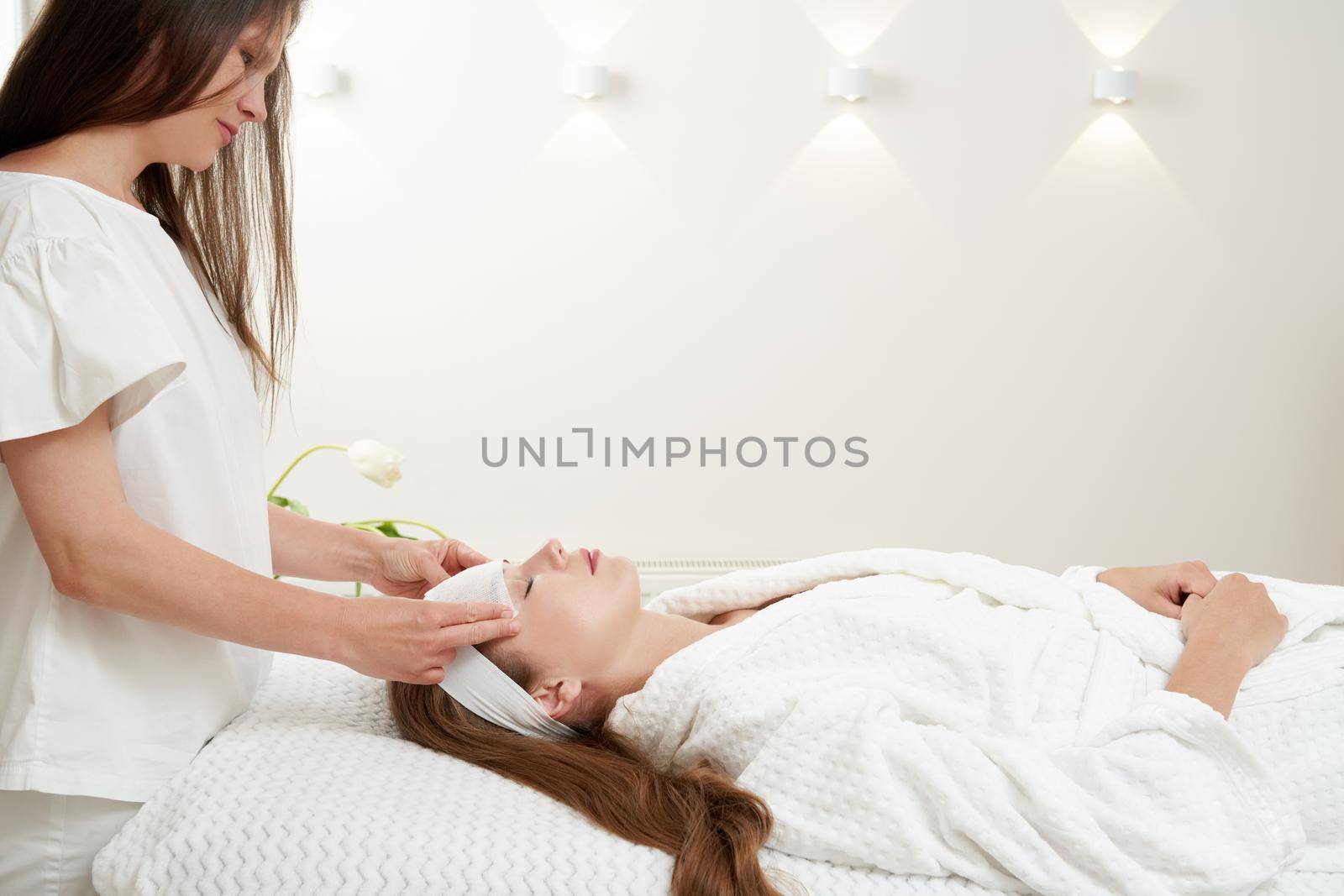 Cosmetologist applying rejuvenating clay facial mask on a female face in beauty salon. Facial treatment concept