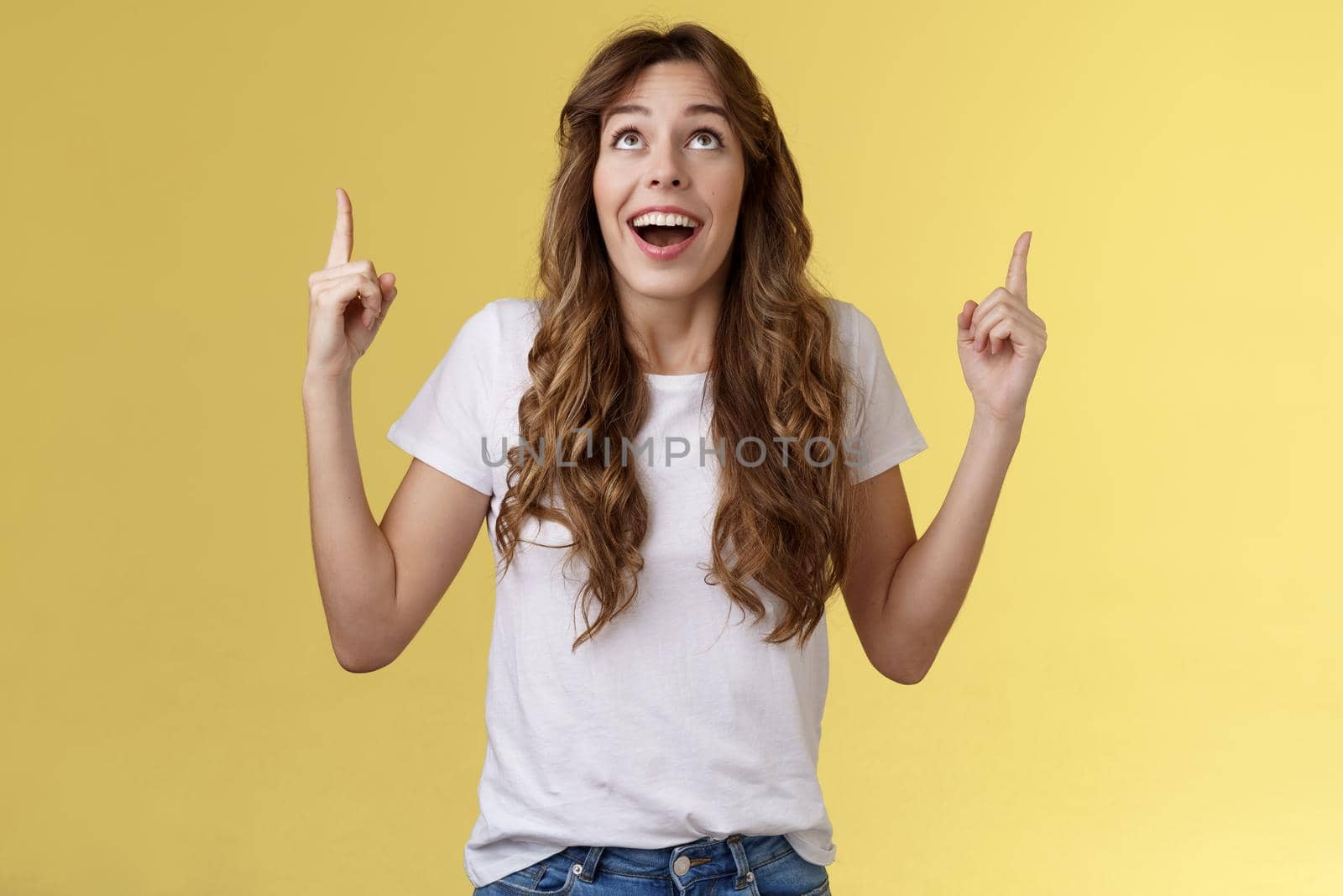 Happy impressed attractive girl long curly haircut drop jaw amused smiling surprised delighted look pointing up index fingers stare admiration fascinated awesome promo offer yellow background.Lifestyle.