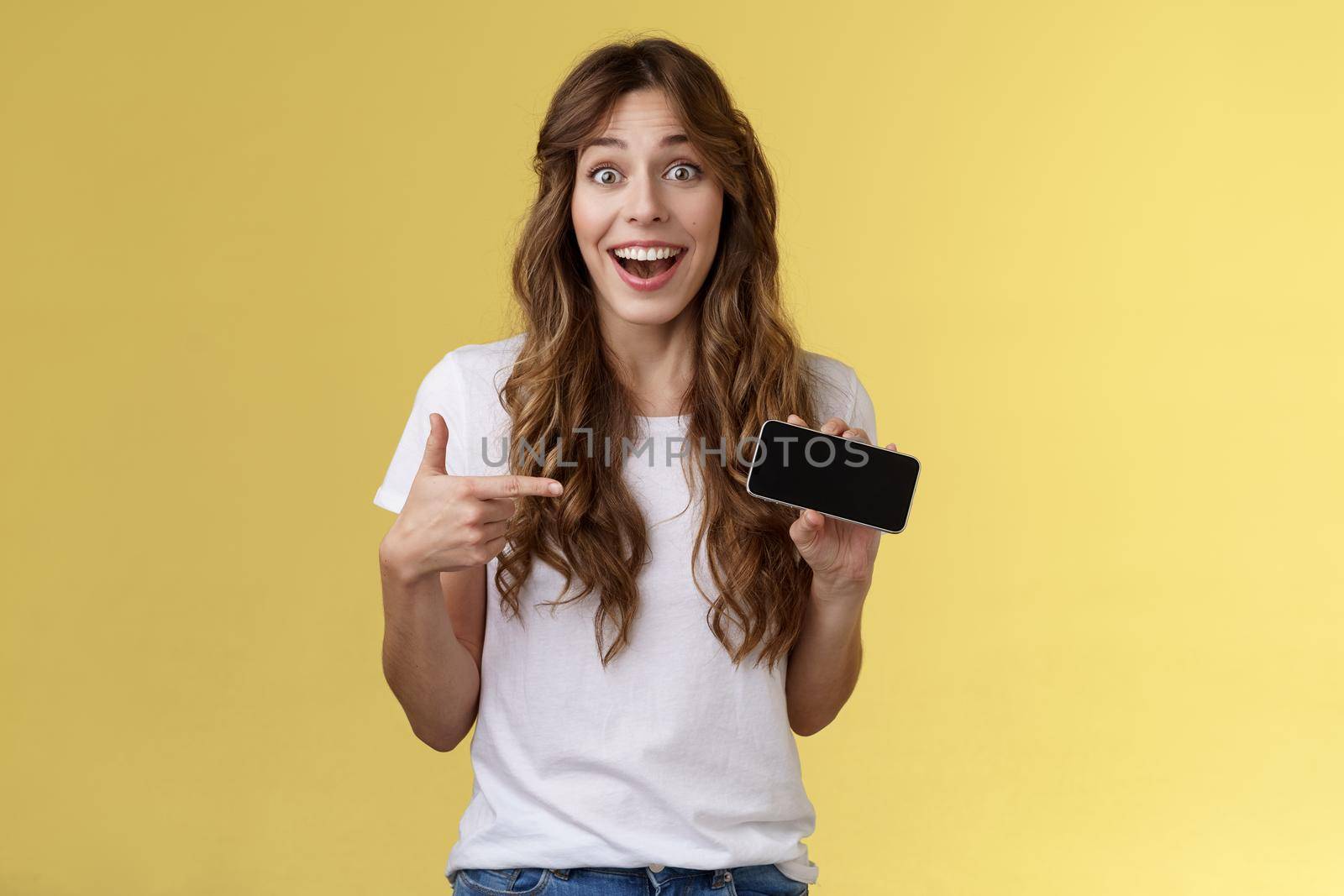 Cheerful surprised cute lucky girl beat best score awesome game showing smartphone display pointing index finger horizontal screen mobile phone introduce cool app smiling broadly yellow background. Lifestyle.