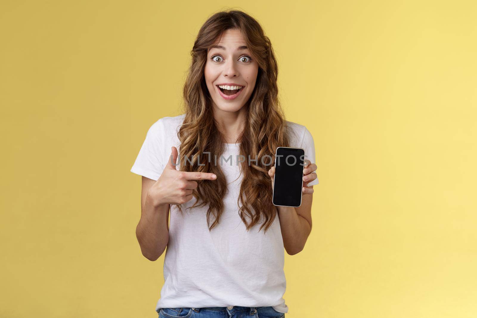 Surprised happy lucky girl winning online internet lottery smiling broadly hold smartphone pointing blank mobile phone screen showing display grinning excited enthusiastic stand yellow background.