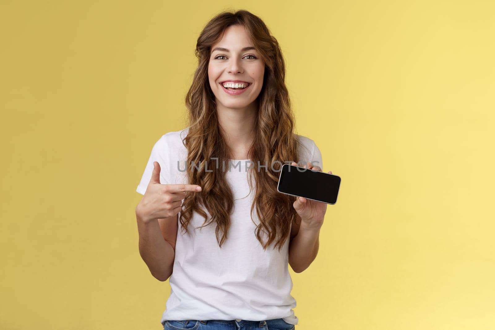 Carefree charismatic cute curly-haired female gamer likes playing smartphone games showing own game score hold mobile phone horizontal pointing display laughing pleased amused. Copy space