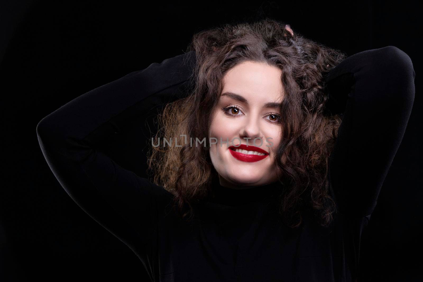 A beautiful brunette girl with curly hair smiles and looks at the camera on a dark background.