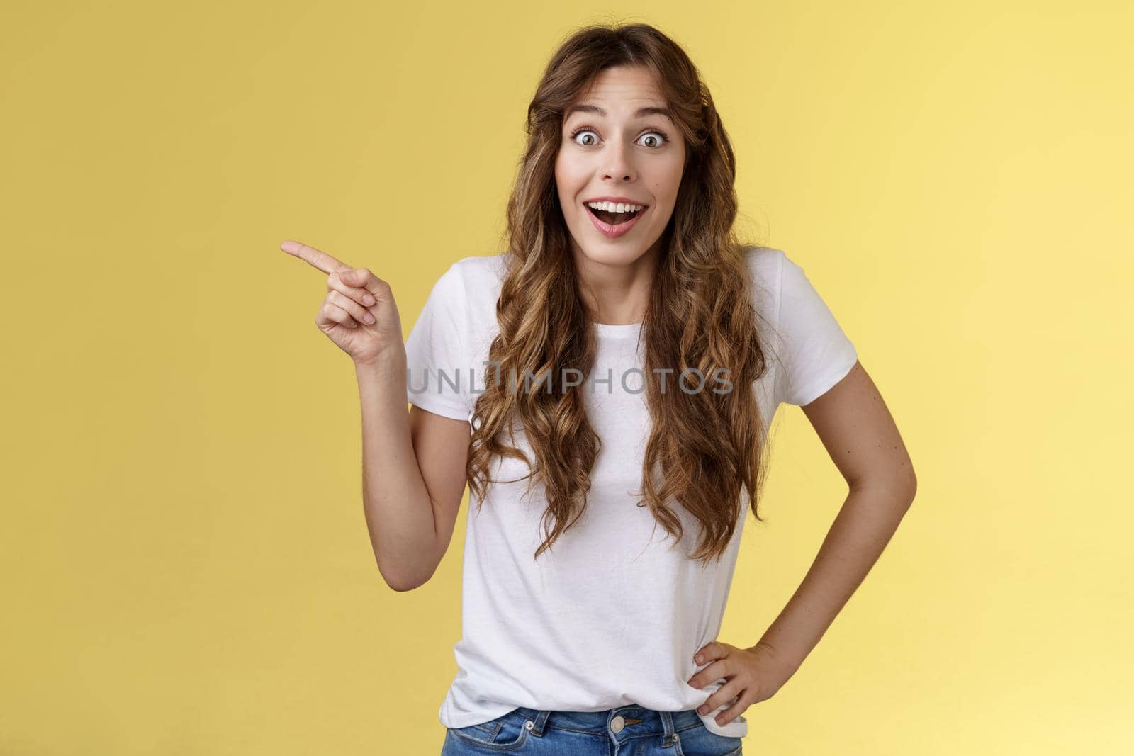 Surprised friendly sociable happy cute girlfriend long curly chestnut haircut smiling impressed gladly pointing left grinning camera toothy astonished expression discuss awesome performance.