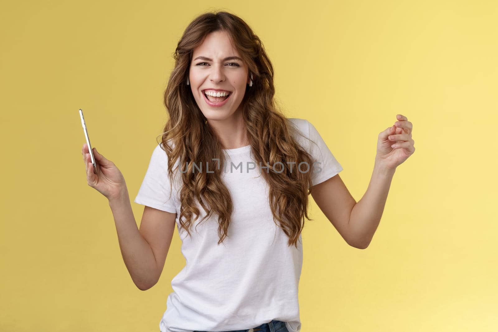 Sassy carefree joyful attractive woman having fun partying listen cool new song favorite artist dancing raise hands up squinting cheeky look camera lip sync hold smartphone yellow background by Benzoix