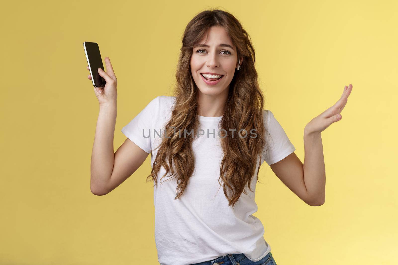 Carefree good-looking lively sociable curly-haired girl having fun wearing wireless earbuds dancing joyfully listening music moving rhythm song hold smartphone raising hands up smiling camera amused by Benzoix