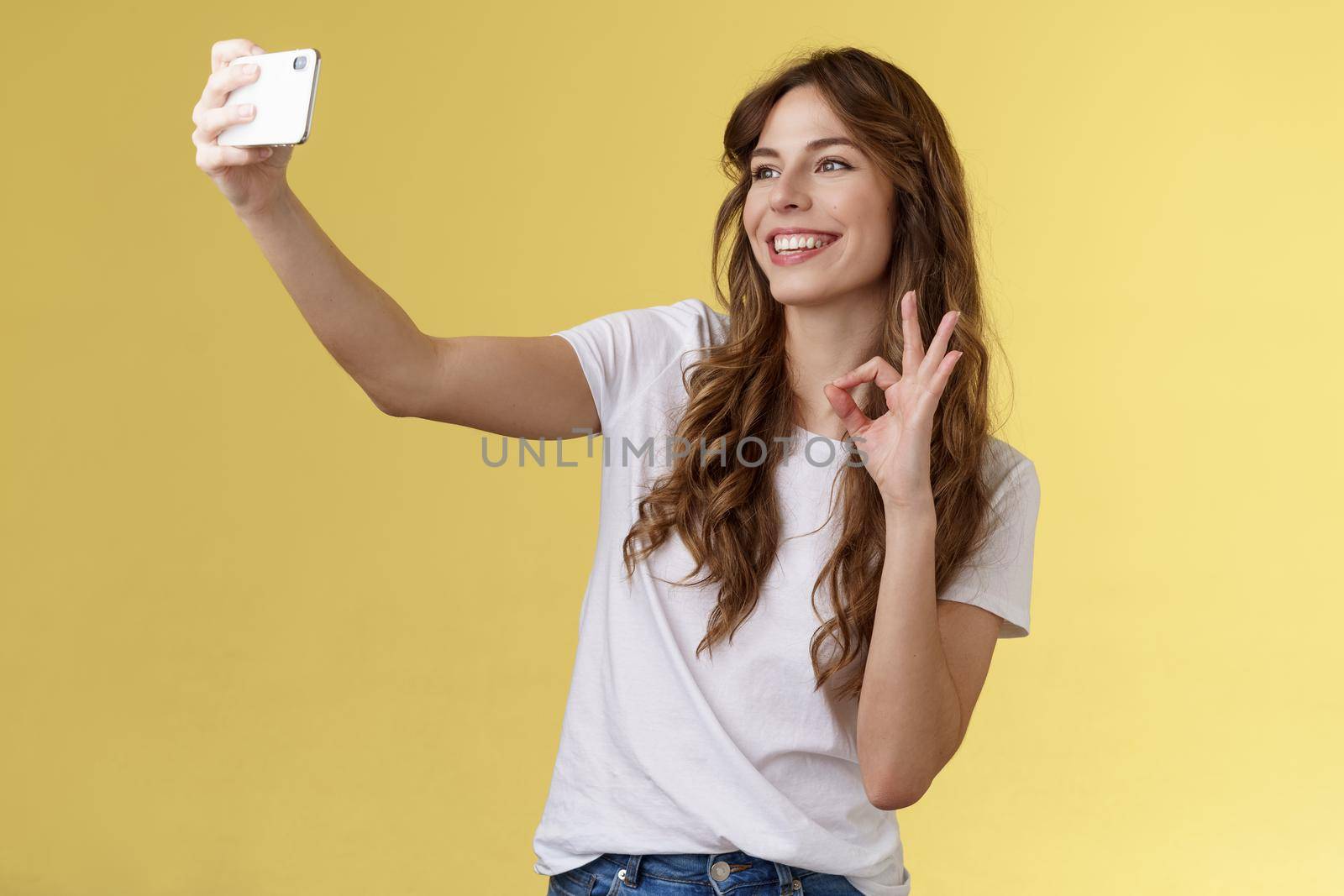 Stylish carefree urban confident curly-haired girl recording video blog followers extend arm hold smartphone taking selfie front camera show okay ok approval gesture yellow background by Benzoix