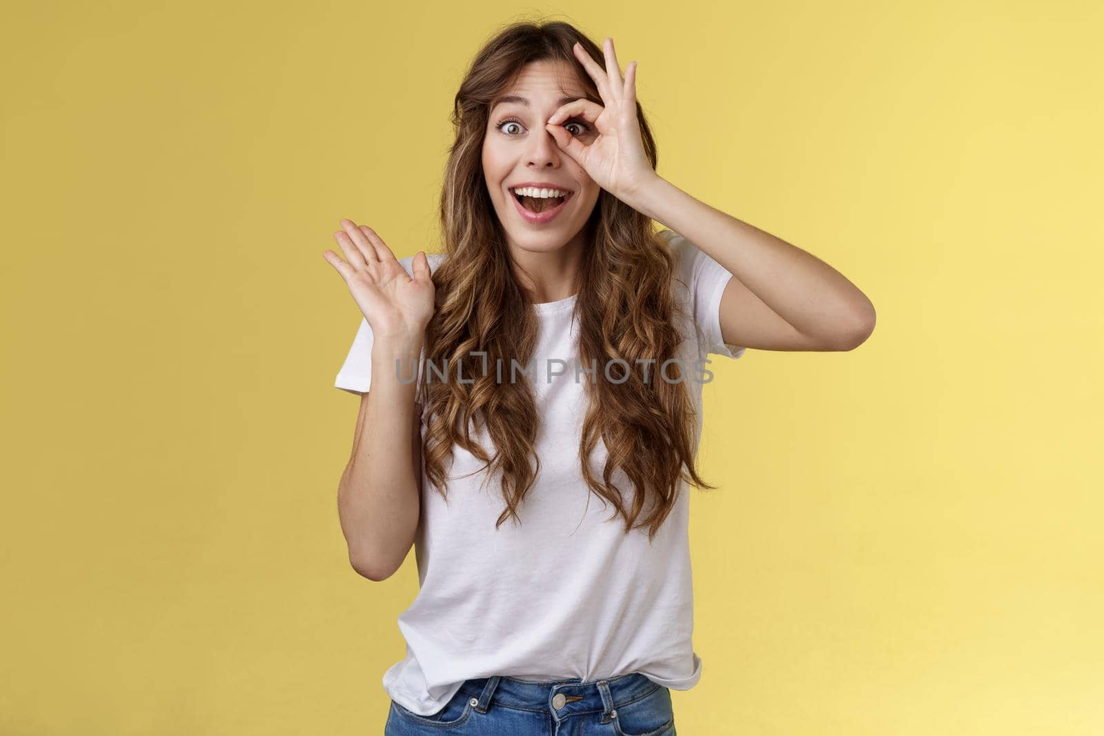 Oh hi I see you. Fascinated cute charming young girl waving palm say hello impressed look through okay ring gesture impressed finally fixed sight stand astonished amazed yellow background.