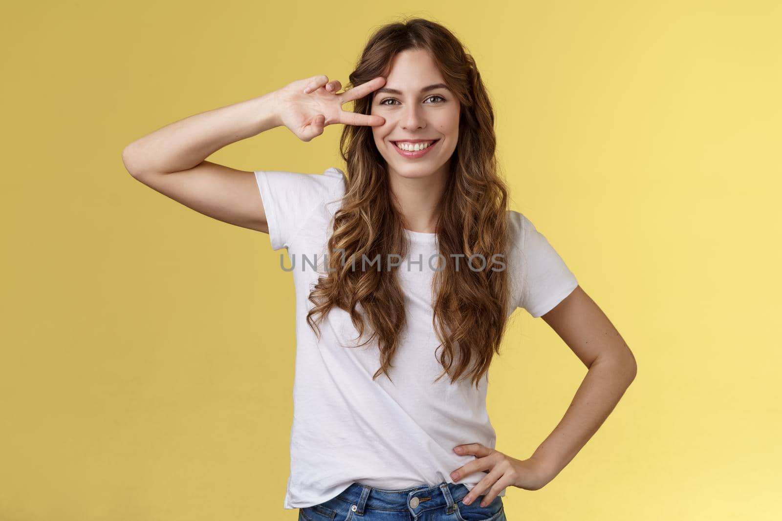 Cheerful positive cute girl curly-haired show peace sign delighted enjoy youth having perfect summer holidays determined rest trip vacation smiling broadly stand confident yellow background. Lifestyle.