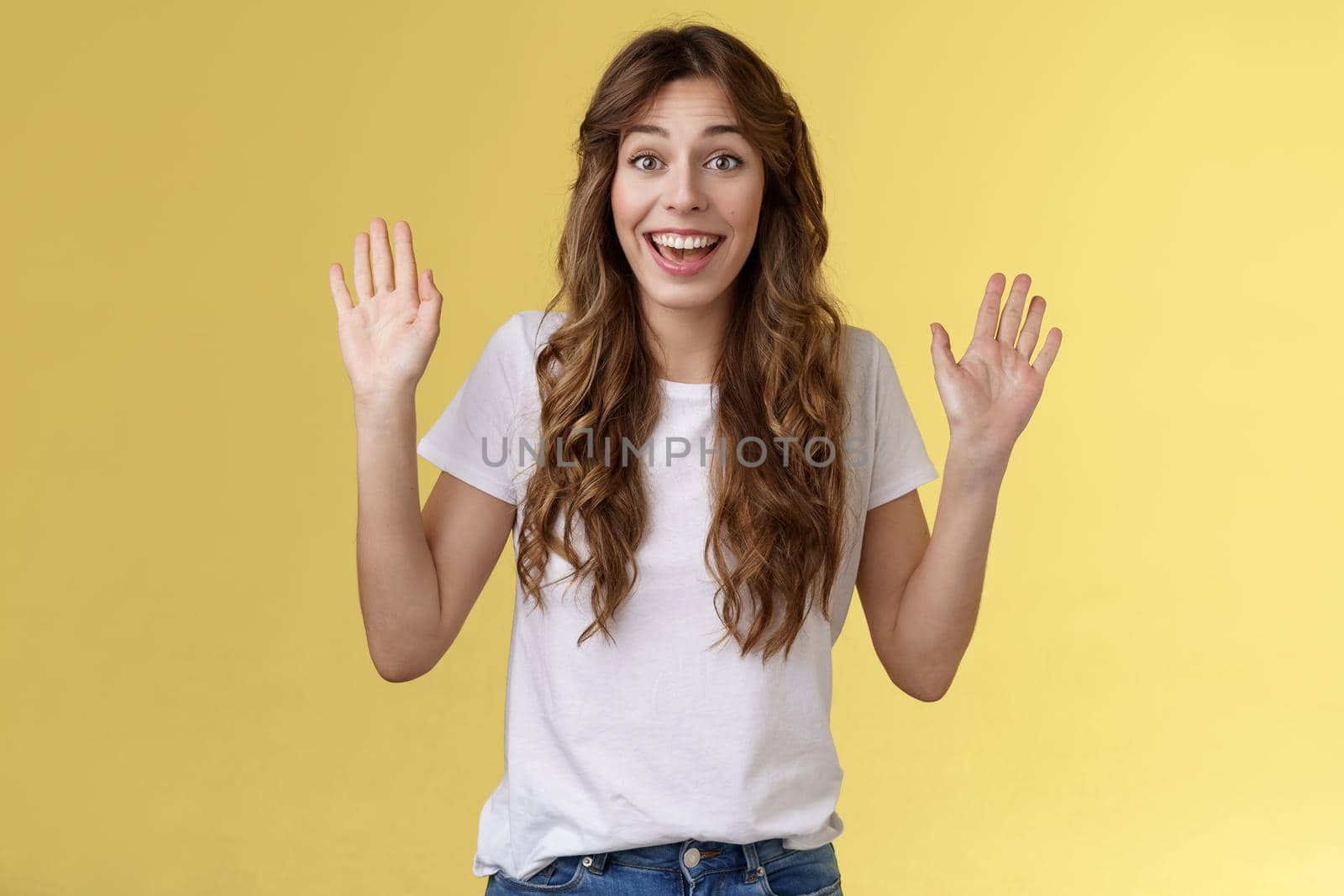 Cheerful glad outgoing cute positive young girl smiling broadly raise both palms waving hands hello greeting gesture welcome friend guest smiling hi gladly invite come inside yellow background. Lifestyle.