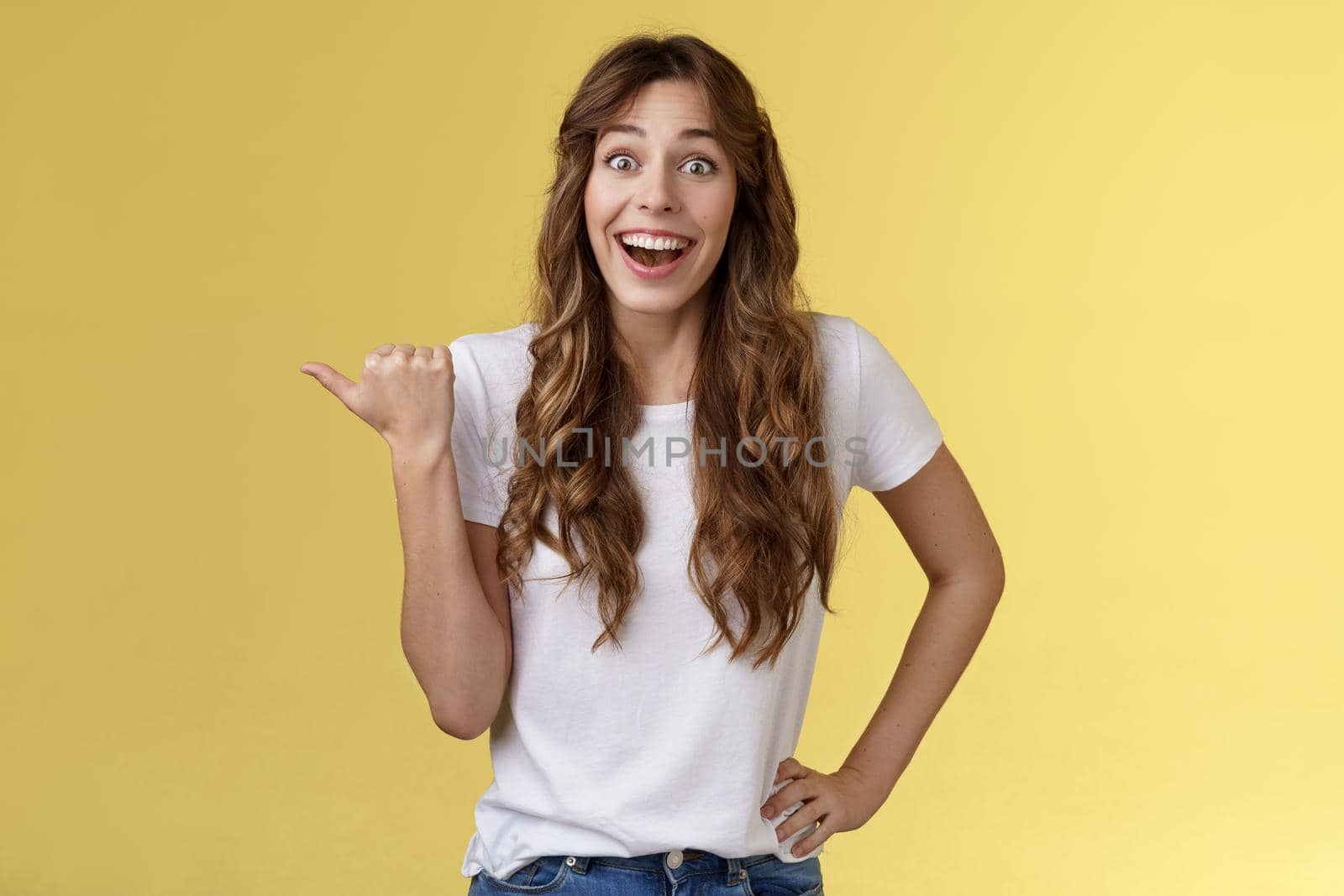 So amazing lets go together. Lively excited european female curly haircut wanna visit cool party smiling joyfully pointing thumb left grinning thrilled look camera admiration yellow background by Benzoix