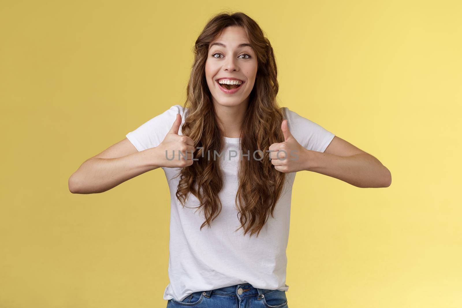 Cheerful suportive energized cute european cute curly-haired girl show thumbs-up approval rooting for friend excellent effort nodding accepting great awesome work done stand yellow background. Lifestyle.