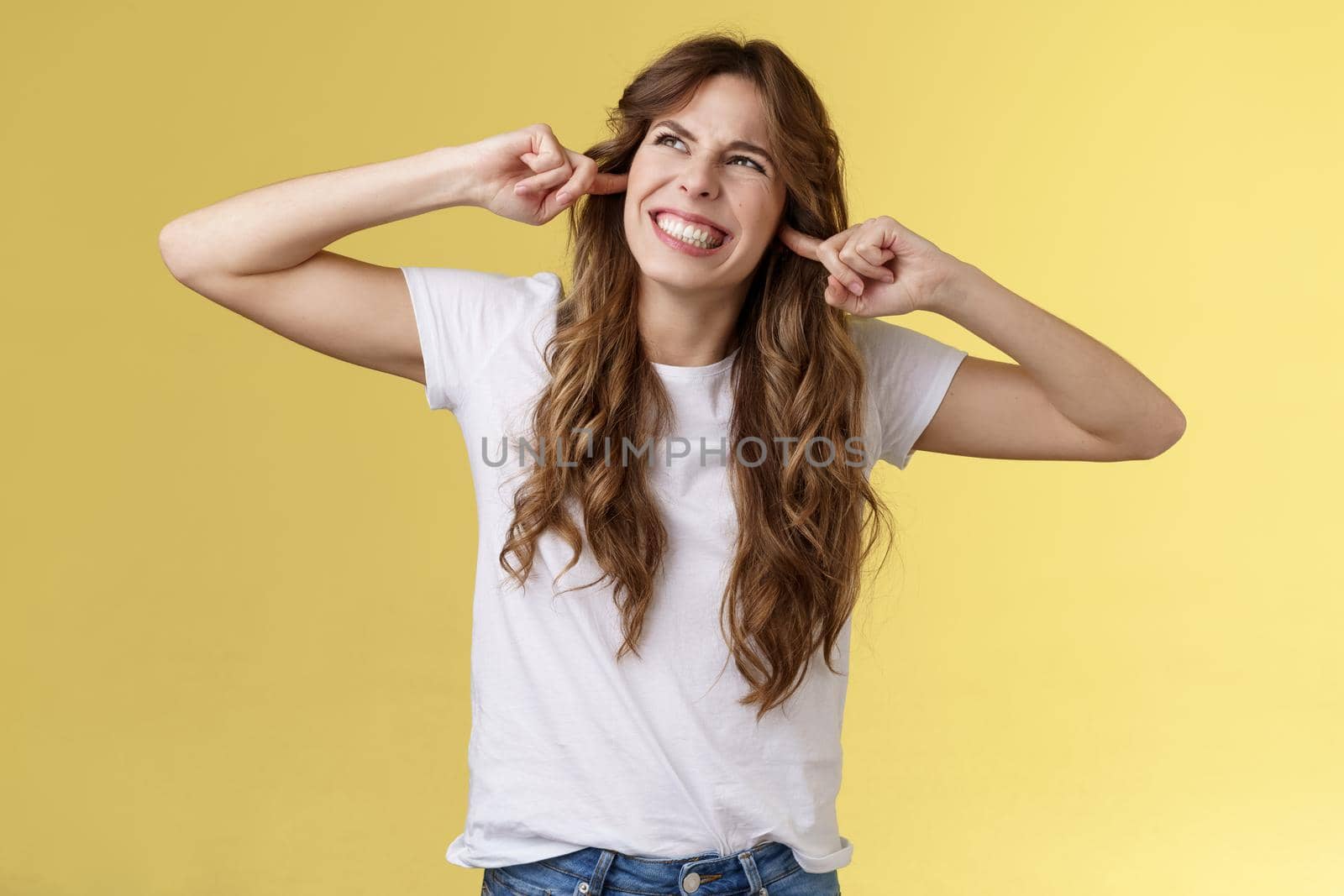 Angry displeased annoyed curly-haired beautiful girl grimacing make wry face stare up irritated noise comming from neighbours above clench teeth plug index fingers ears hate loud music playing by Benzoix