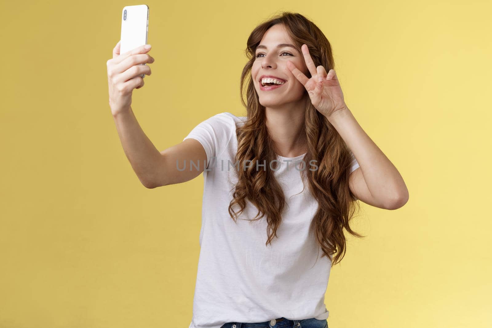 Positive charisamtic happy young girl curly-haired raise smartphone taking selfie show peace victory sign having video call stand yellow background smiling mobile front camera posing lively. Lifestyle.