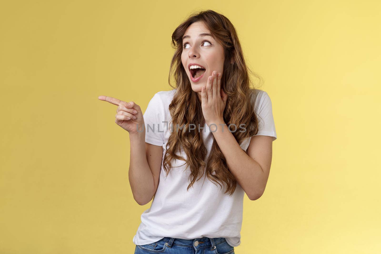 Silly flirty young attractive woman sighing admiration excitement checking out amazing stunning promo drop jaw smiling touch cheek touched delighted turn pointing left amused yellow background. Lifestyle.