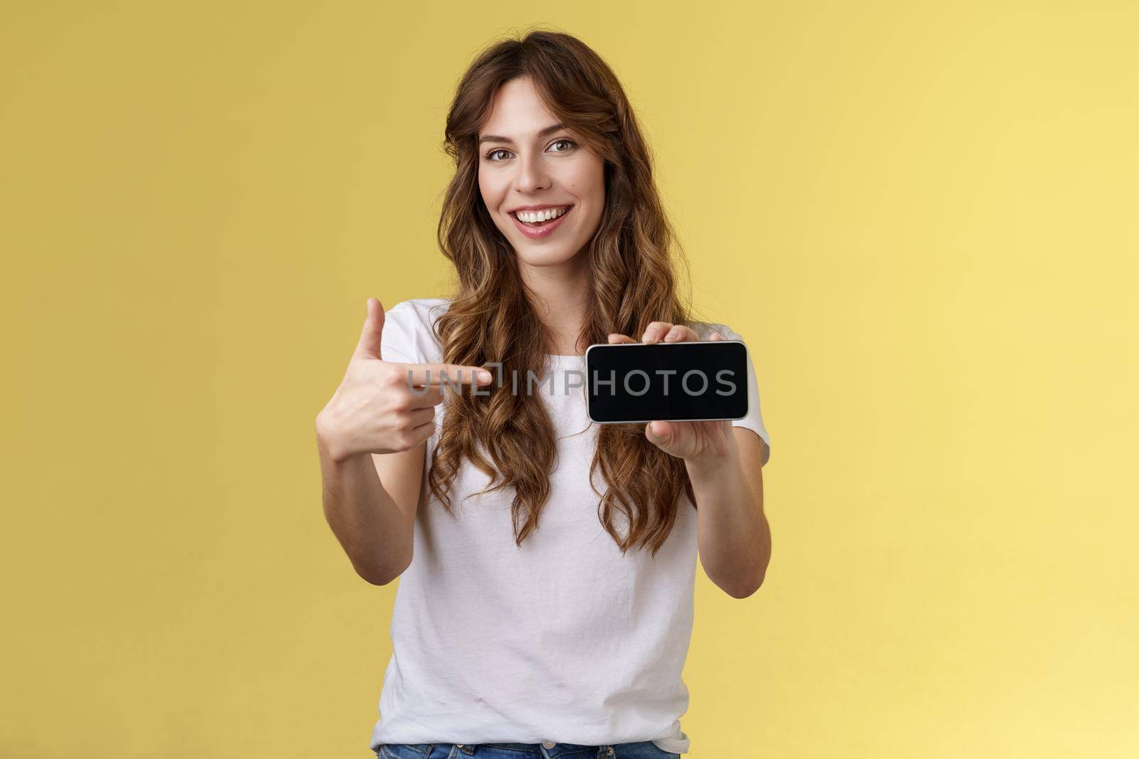 Confident sassy good-looking stylish woman introduce awesome game app show smartphone pointing index finger blank mobile phone screen smiling delighted recommend try application yellow background by Benzoix