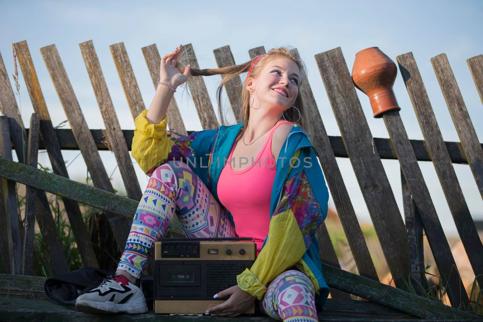 A beautiful country girl in bright clothes sits on a wooden bench with a cassette recorder. Woman in the style of the 90s.