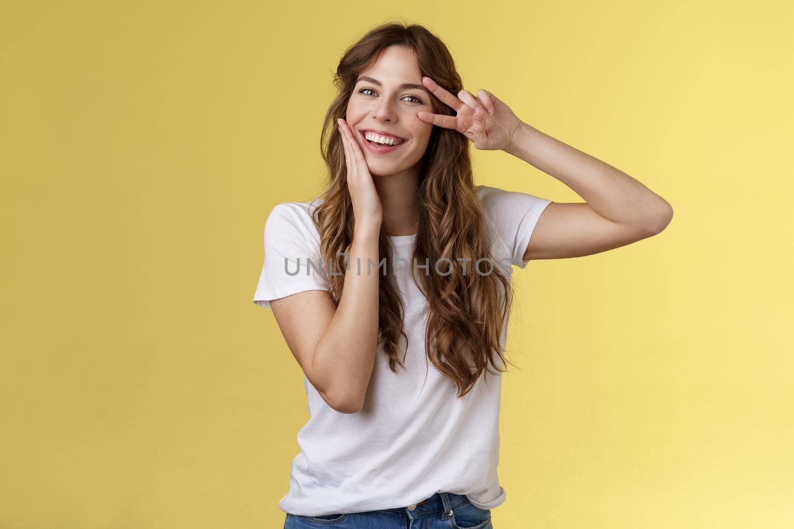 Cheerful positive lovely girlfriend curly hairstyle touch cheek blushing modest cute showing victory peace sign near eye optimistic upbeat attitude having fun enjoying summer yellow background. Lifestyle.