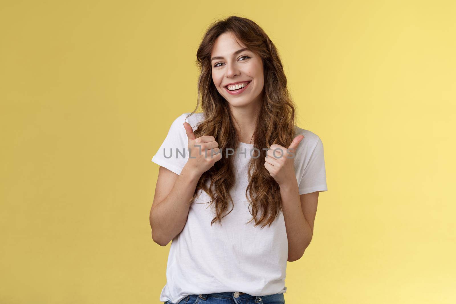 Good have my support. Cheerful positive cute lively curly-haired european girl show thumbs-up approval like gesture tilt head pleased accepting judging fine choice stand yellow background.