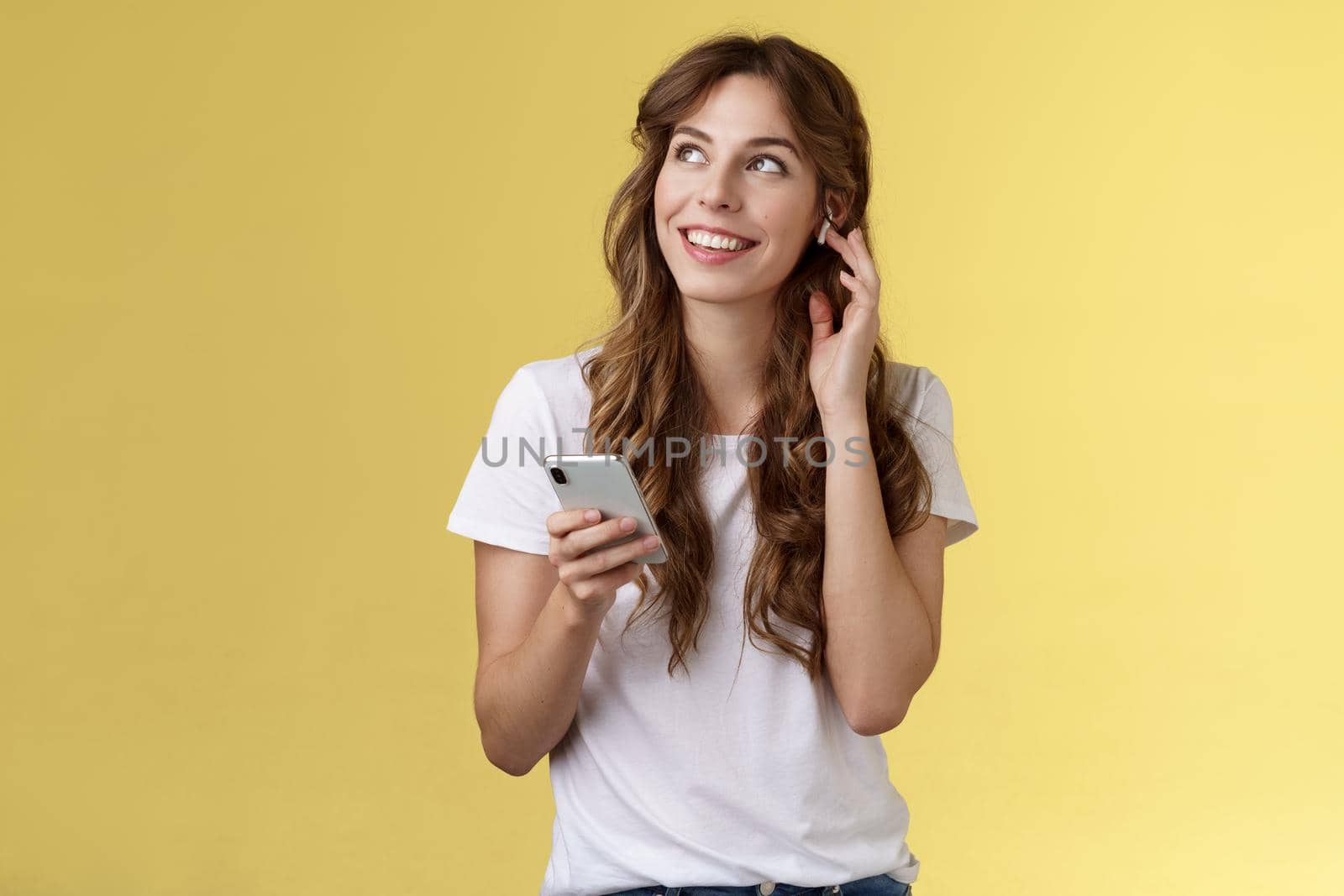 Dreamy happy cheerful curly-haired girl look around contemplate beautiful summer weather listening music touch wireless earbud calling friend talking via earphones hold smartphone yellow background. Lifestyle.