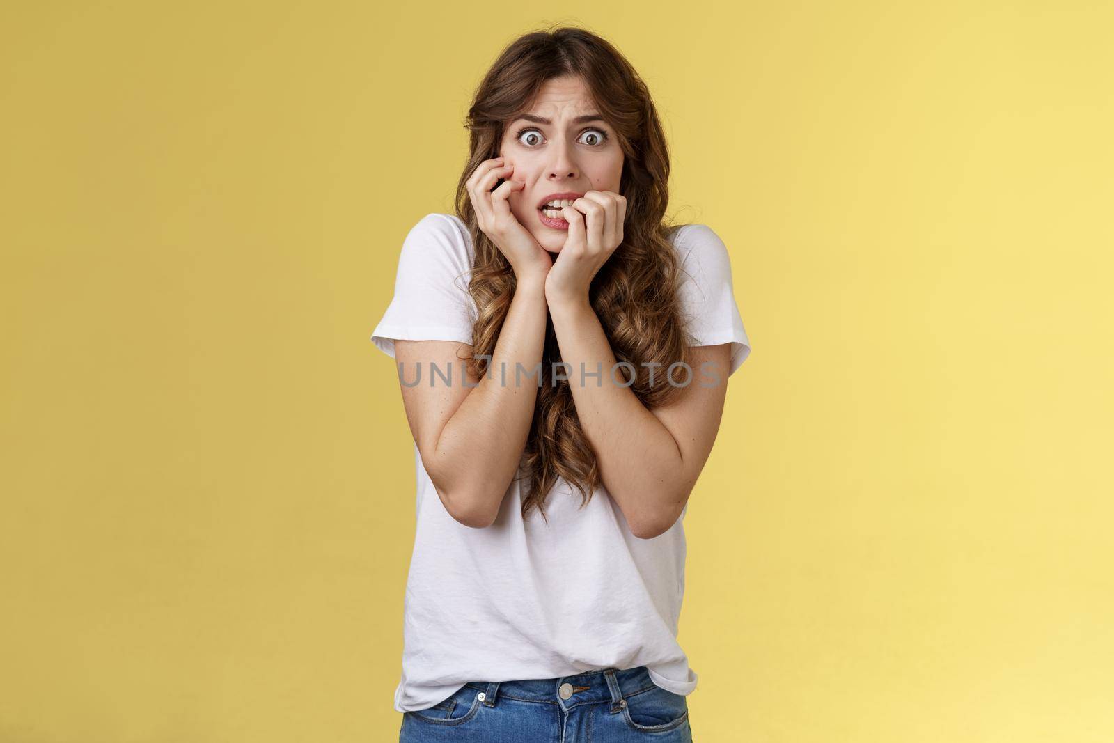 Girl panicking feeling scared timid insecure biting fingernails stare camera frightened clench teeth trembling fear touch face nervously feeling unconfident terrified stand yellow background.