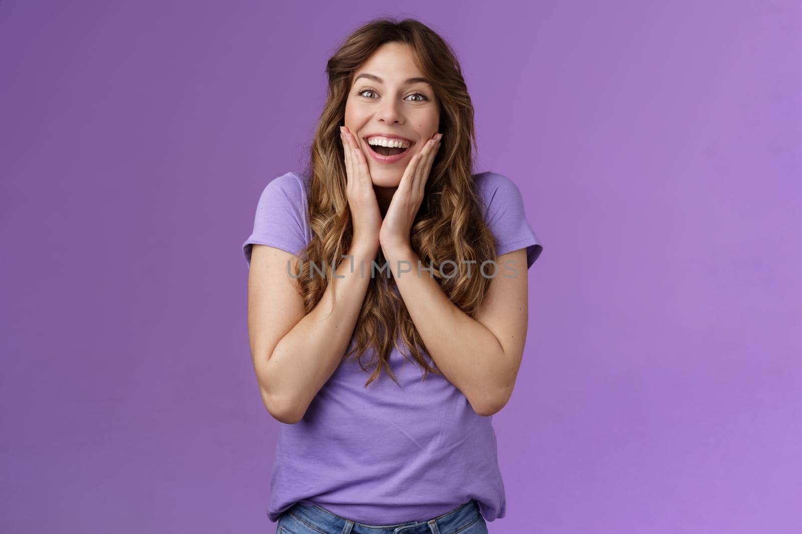 Happy lively lucky enthusiastic girl curly hairstyle open mouth admiration fascinated smiling broadly touch cheek impressed surprised awesome incredible positive news stand purple background.