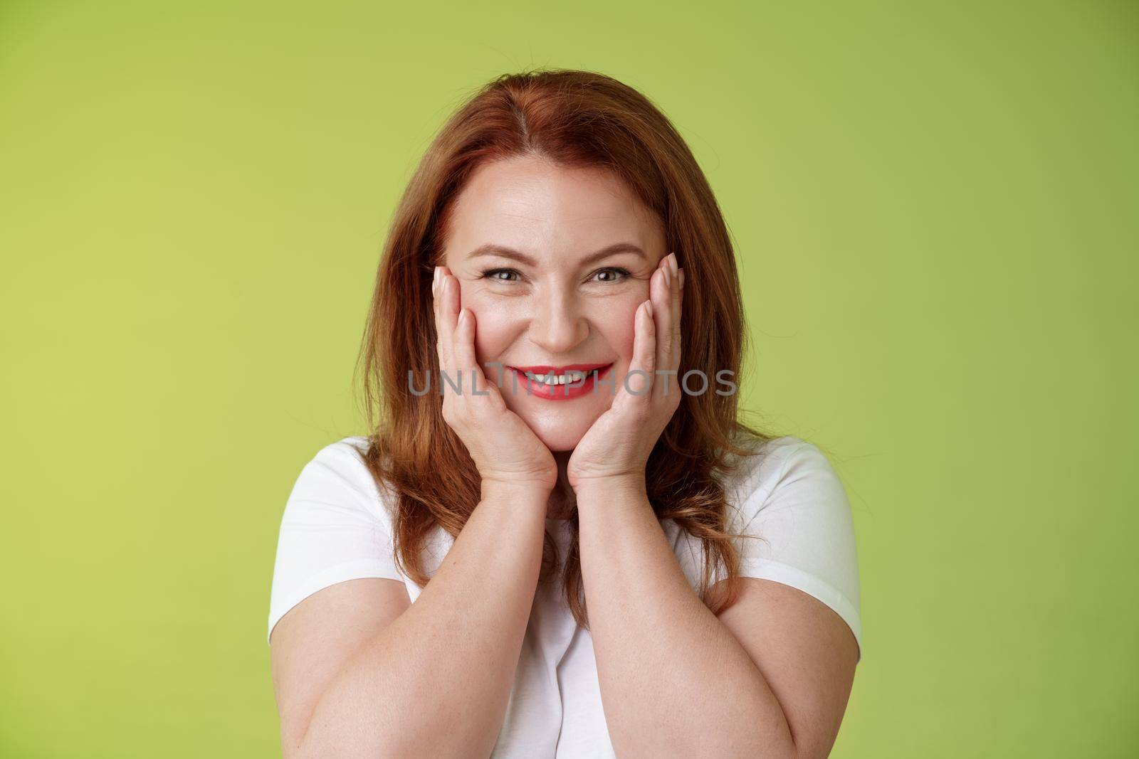 Happy delighted cheerful lucky redhead middle-aged caucasian woman blushing joyfully receive touching cute gift touch cheeks pleased delighted smiling broadly feel happiness joy green background by Benzoix