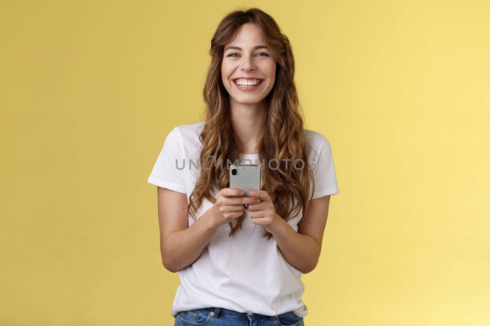 Cheerful carefree happy lively girl hold smartphone using awesome funny app playing cool game relaxing waiting friend park browsing social media using mobile phone look camera upbeat smiling by Benzoix