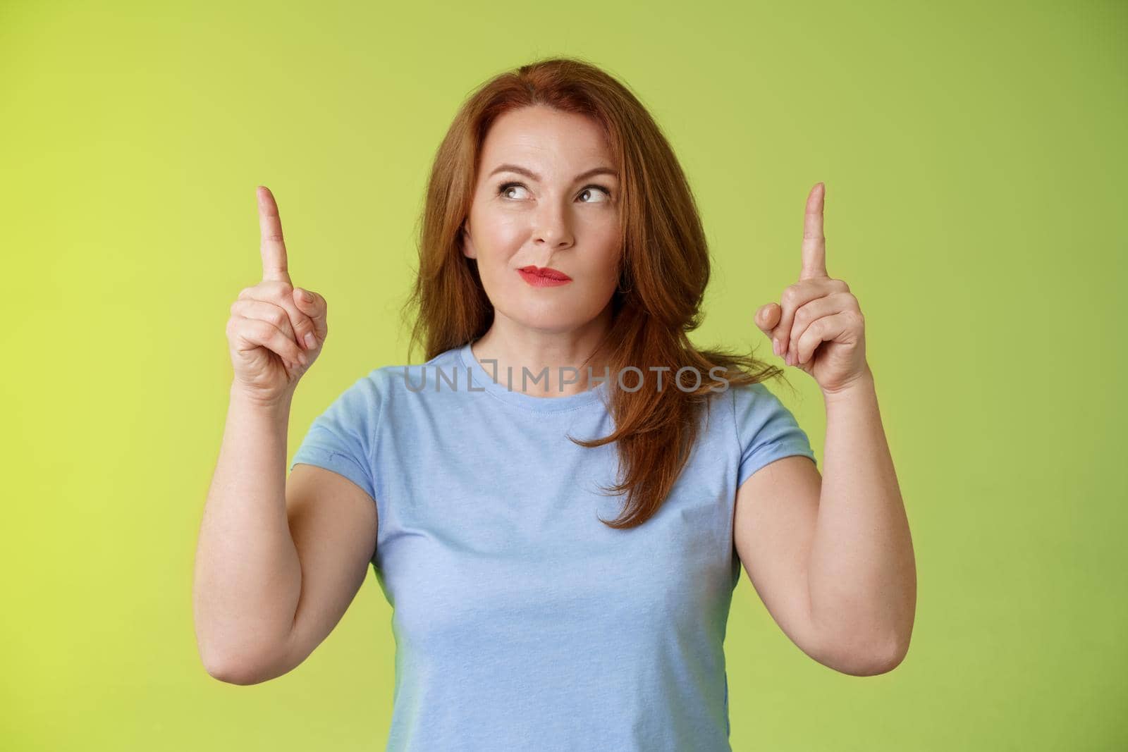 Curious thoughtful cute redhead female middle-aged ginger mother smirking thinking look aside pointing up index fingers deciding choice pondering choosing between variants green background.